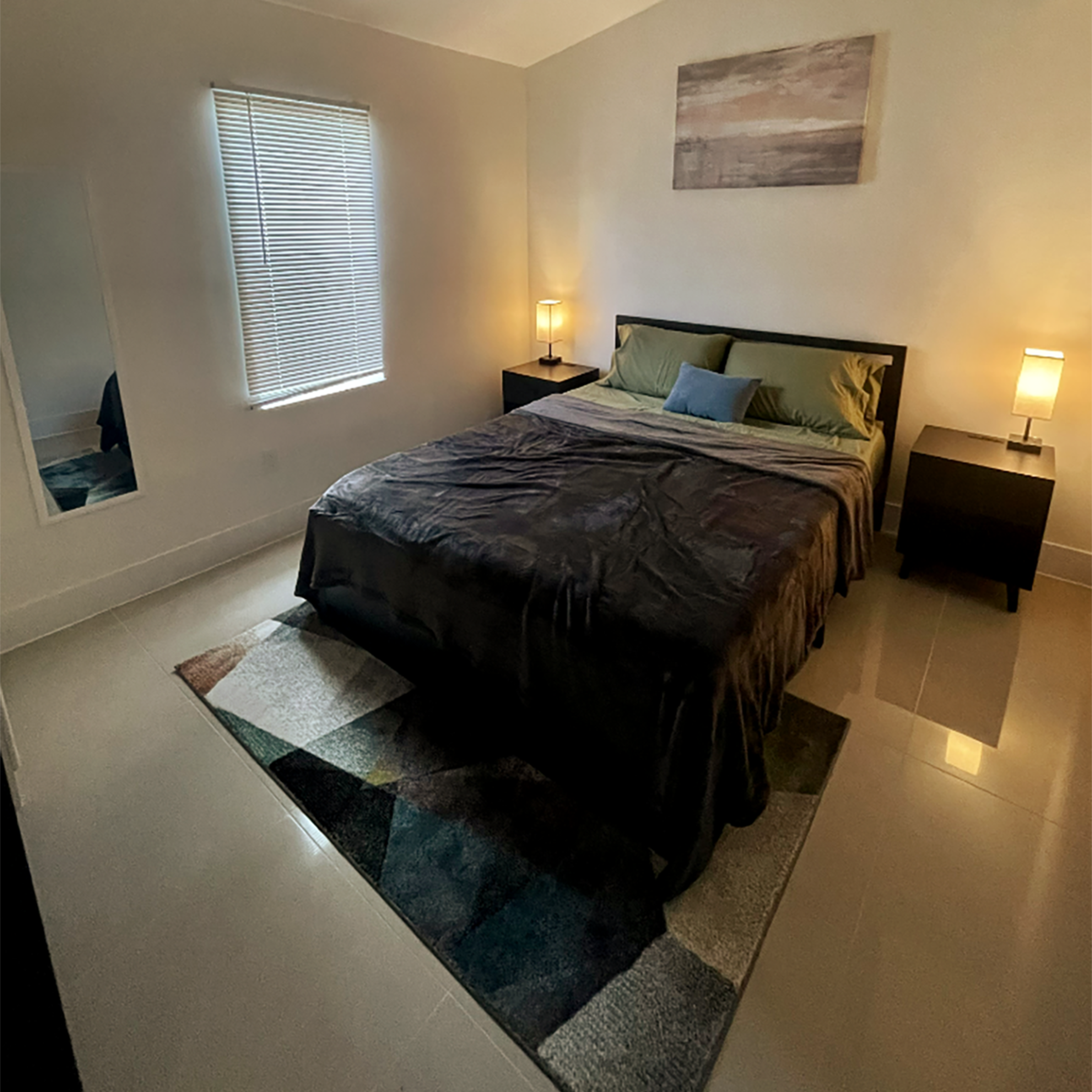 A bedroom with a bed, nightstands, lamps, a large mirror, and a colorful rug.