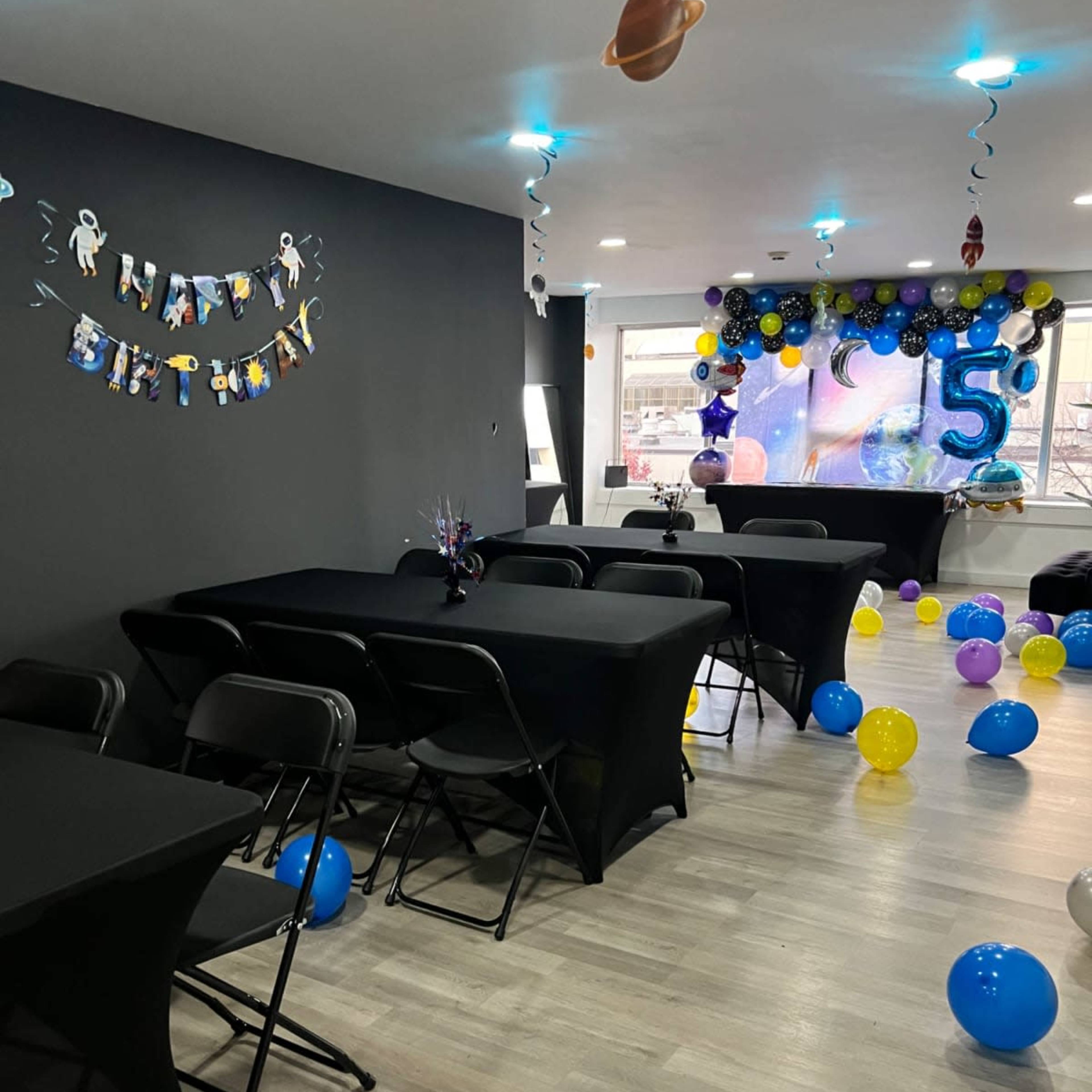 A party room decorated for a birthday celebration, featuring black tables, colorful balloons, and a "Happy Birthday" banner on the wall.