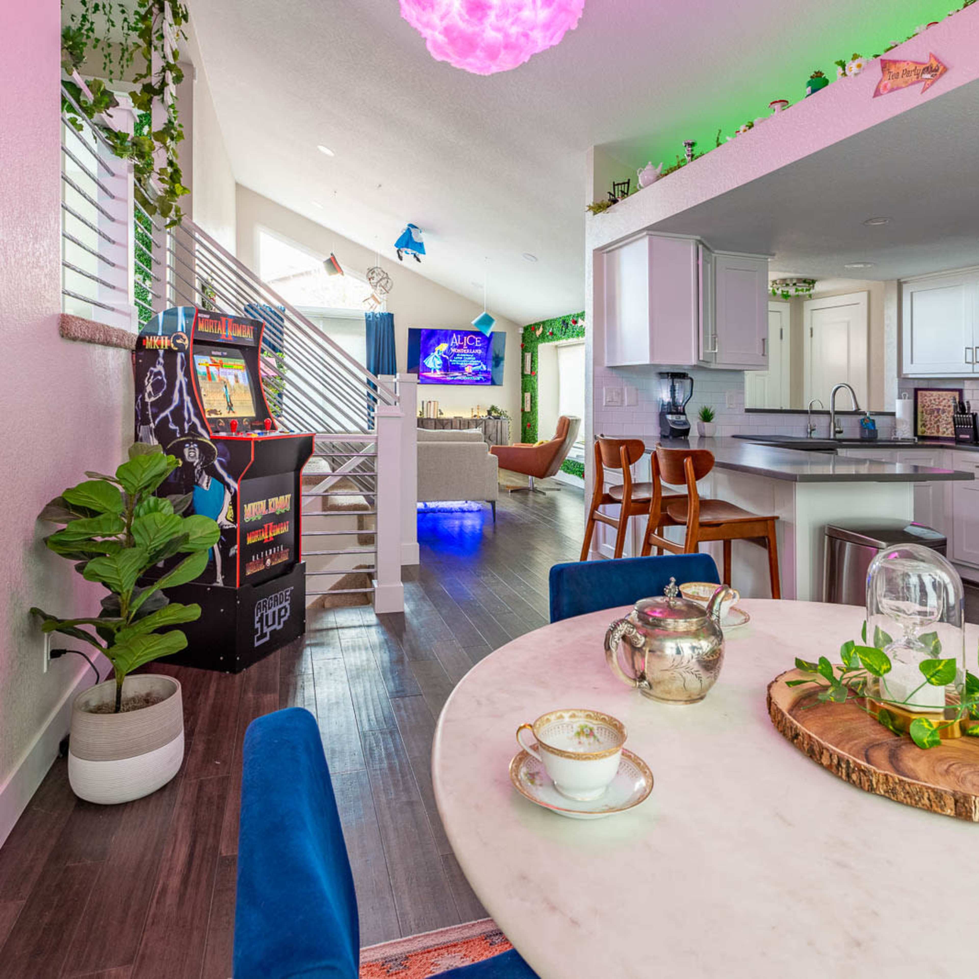 A bright, modern kitchen and dining area features a marble table with tea cups, an arcade game, and a view of a living room decorated with plants and colorful lighting.