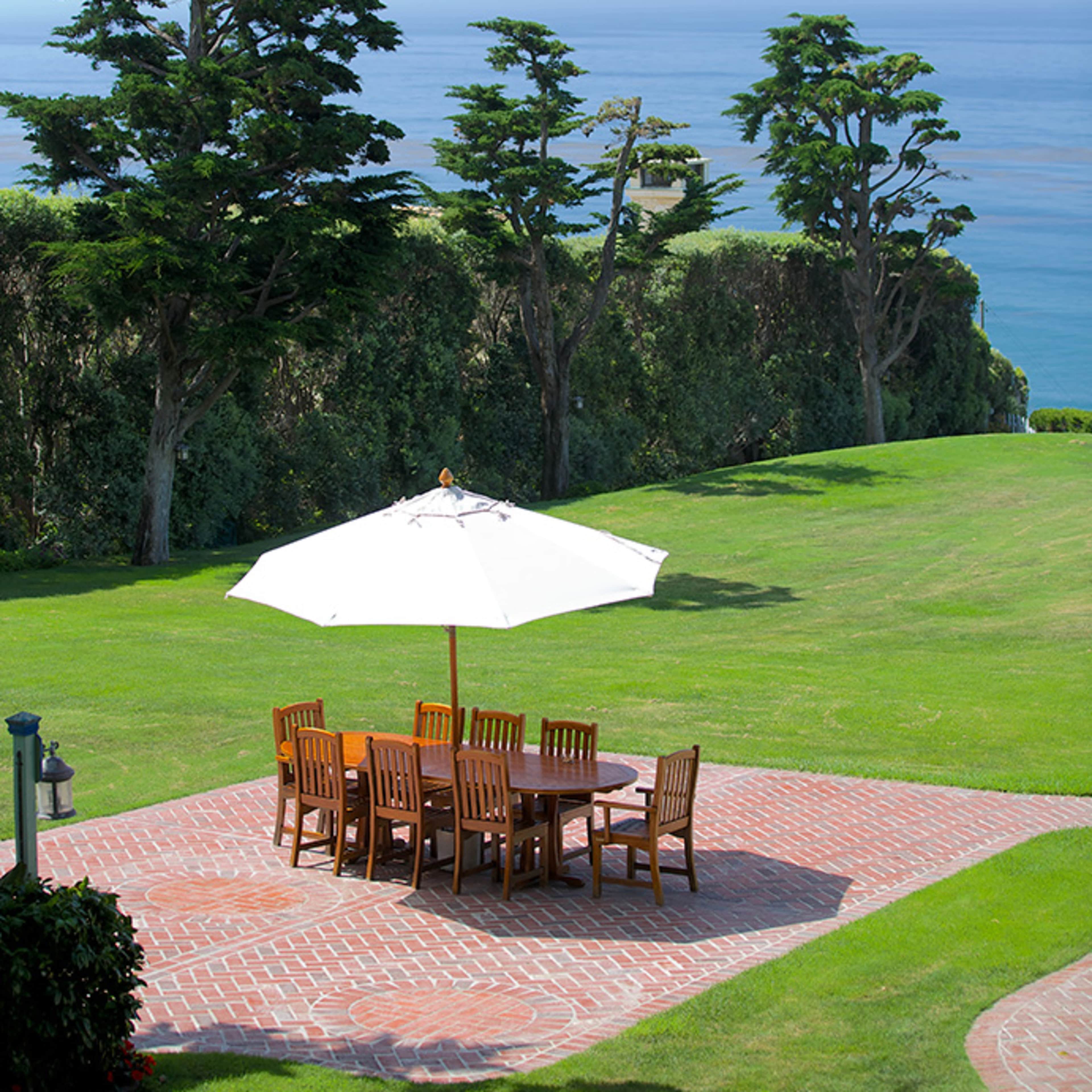 A patio with a large table and chairs is positioned under a white umbrella overlooking the ocean.