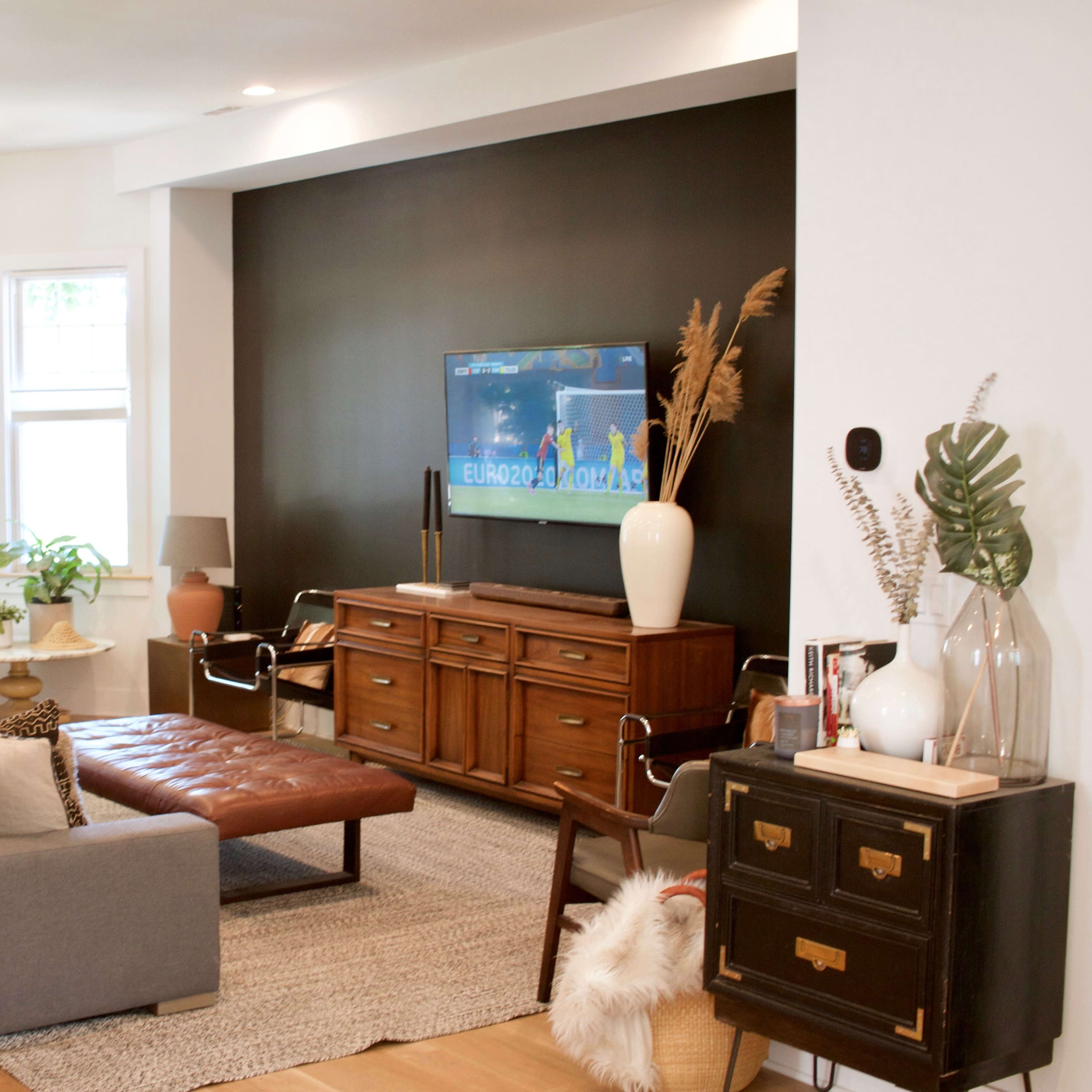 The living room features a gray sectional sofa, a leather coffee table, and a dark accent wall with a television, surrounded by large windows and decorative plants.