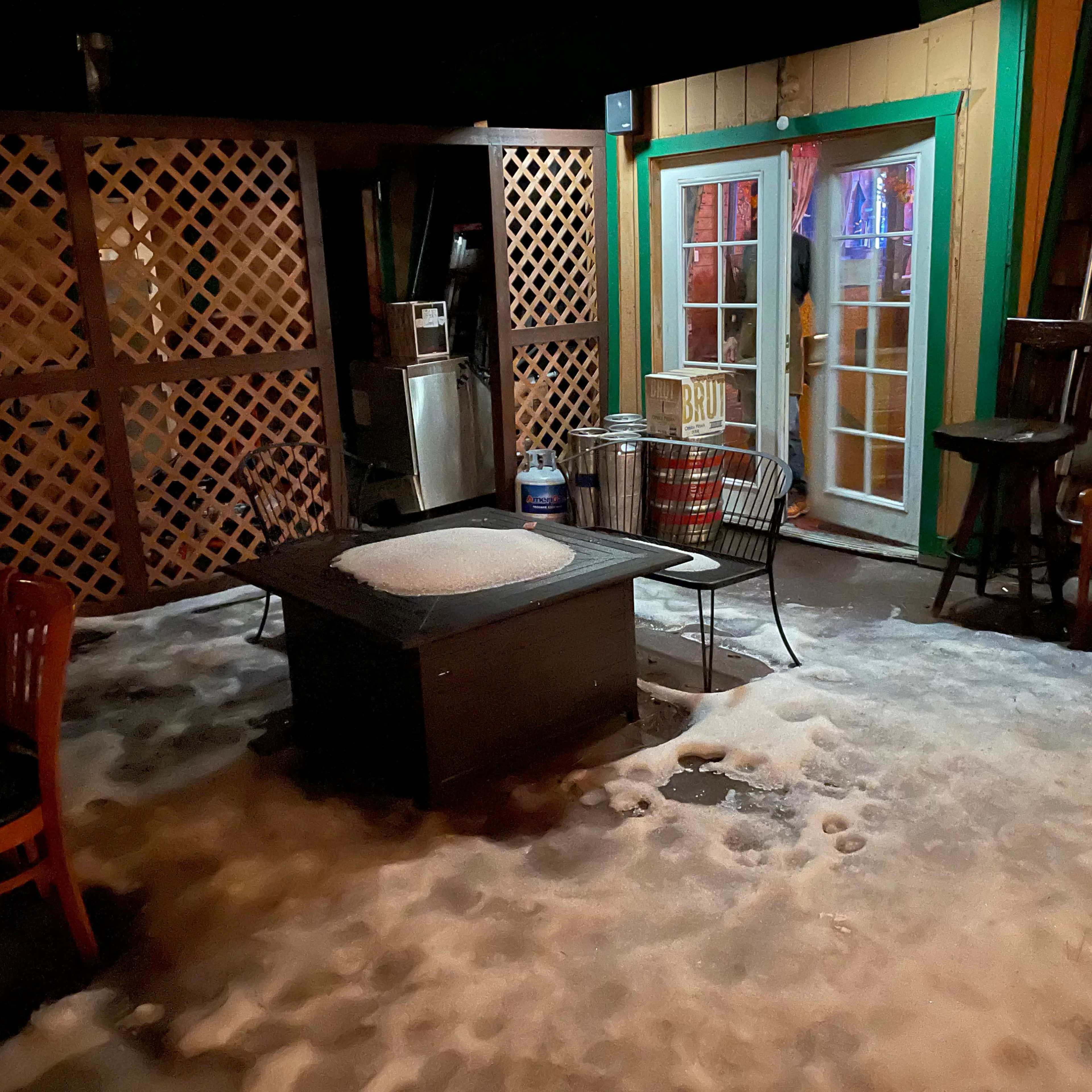 A snowy outdoor patio features scattered chairs, a table with a circular snow mound, and a lit door in the background.