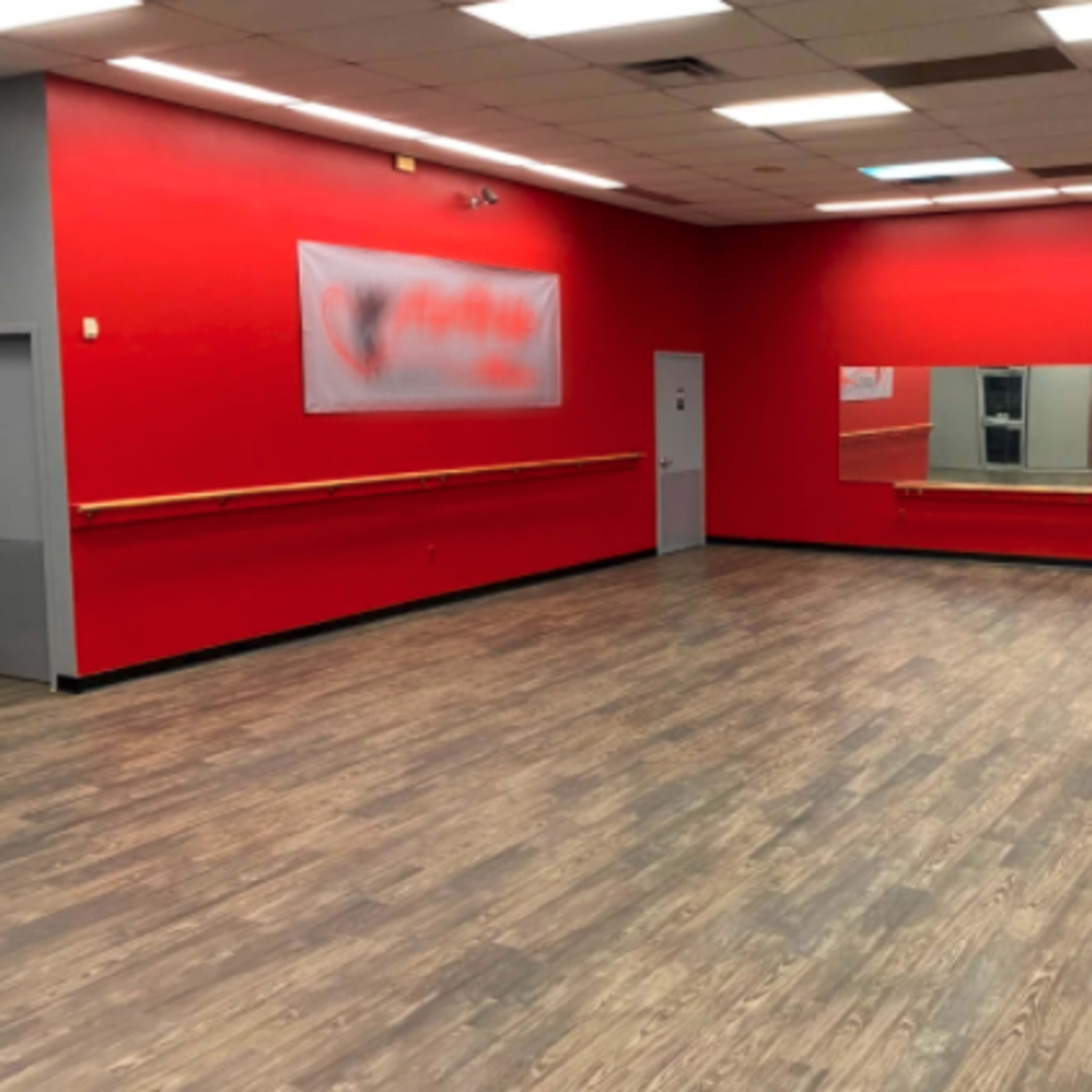 An empty dance studio with red walls, a wooden floor, a long mirror on one side, and a barre along the wall.