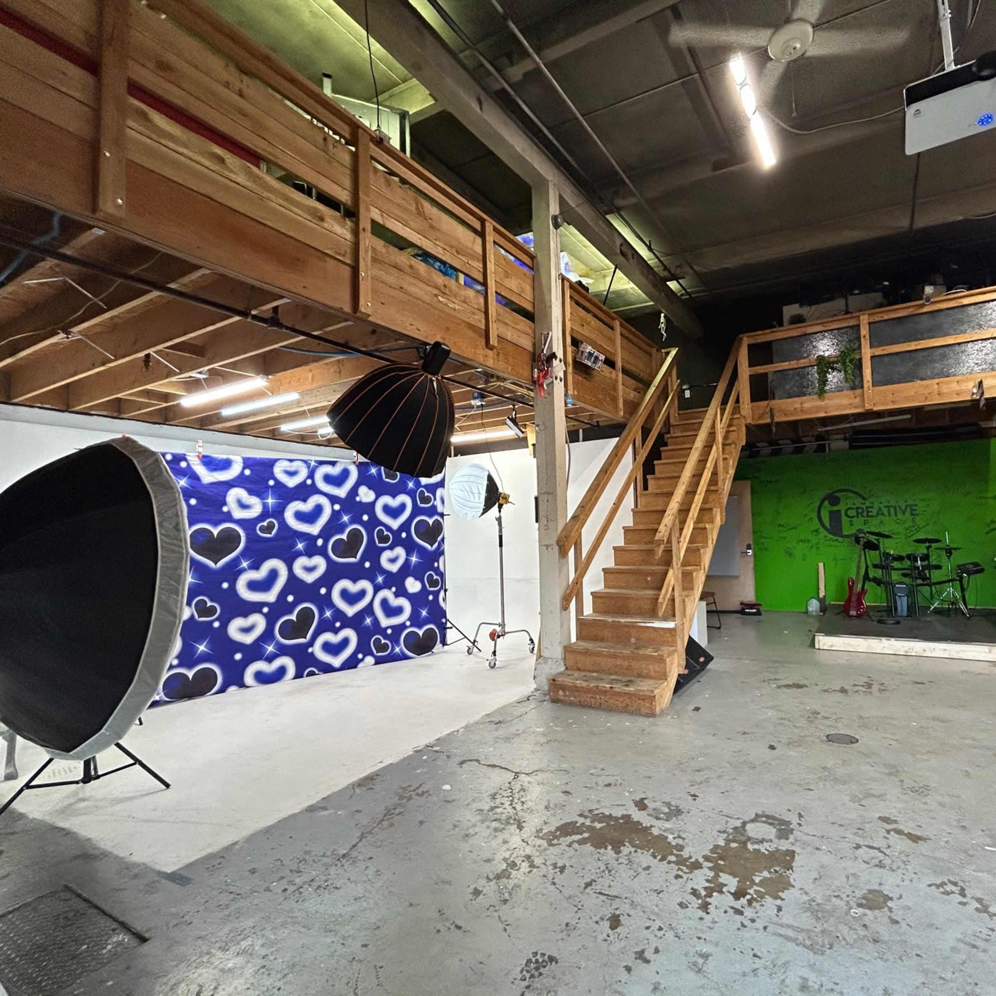 A spacious studio with a staircase leading to a loft area, equipped with photography backdrops and lighting equipment, as well as a green wall featuring the word "Creative."