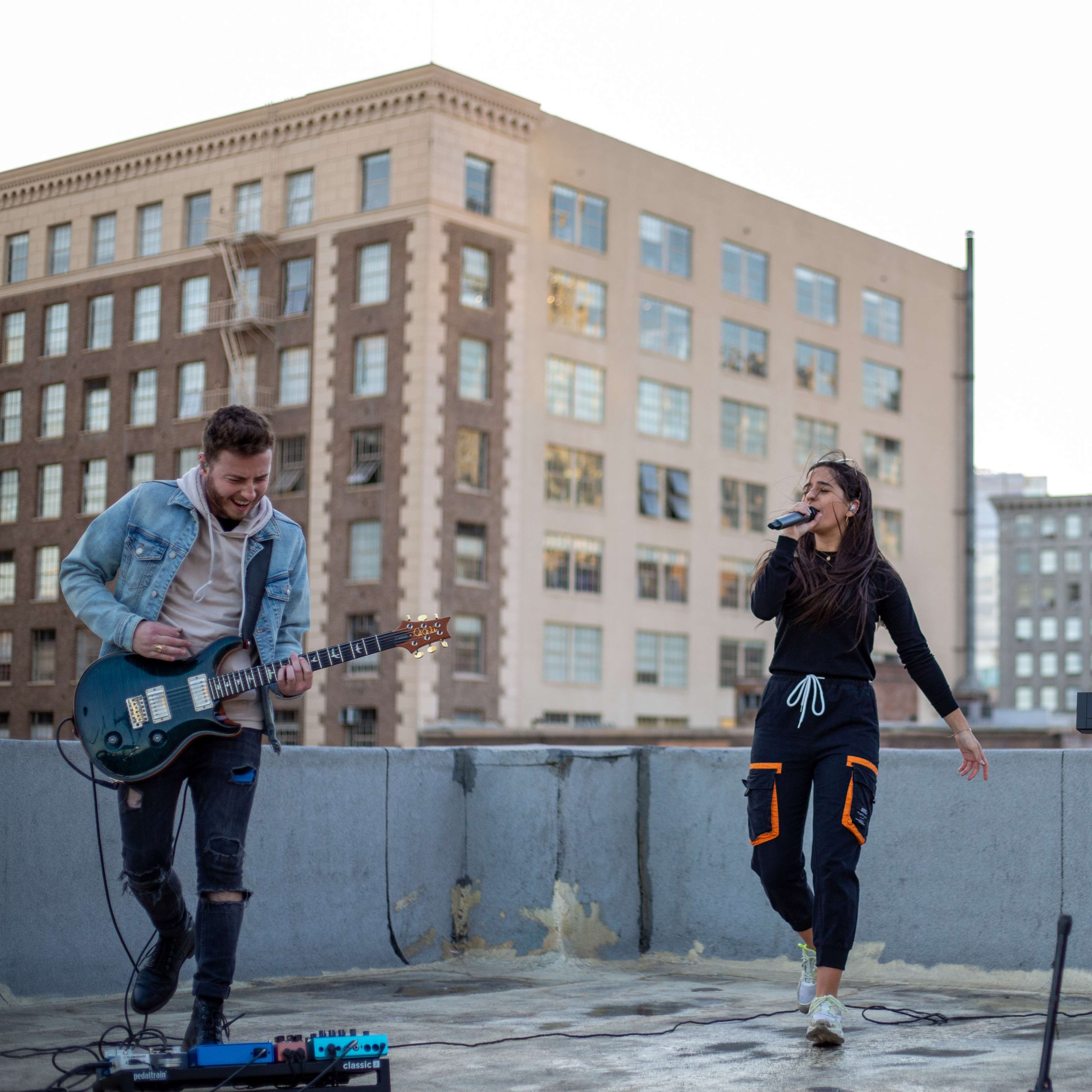 Three musicians perform on a rooftop, with one playing guitar, one singing, and another playing a keyboard against a backdrop of city buildings.