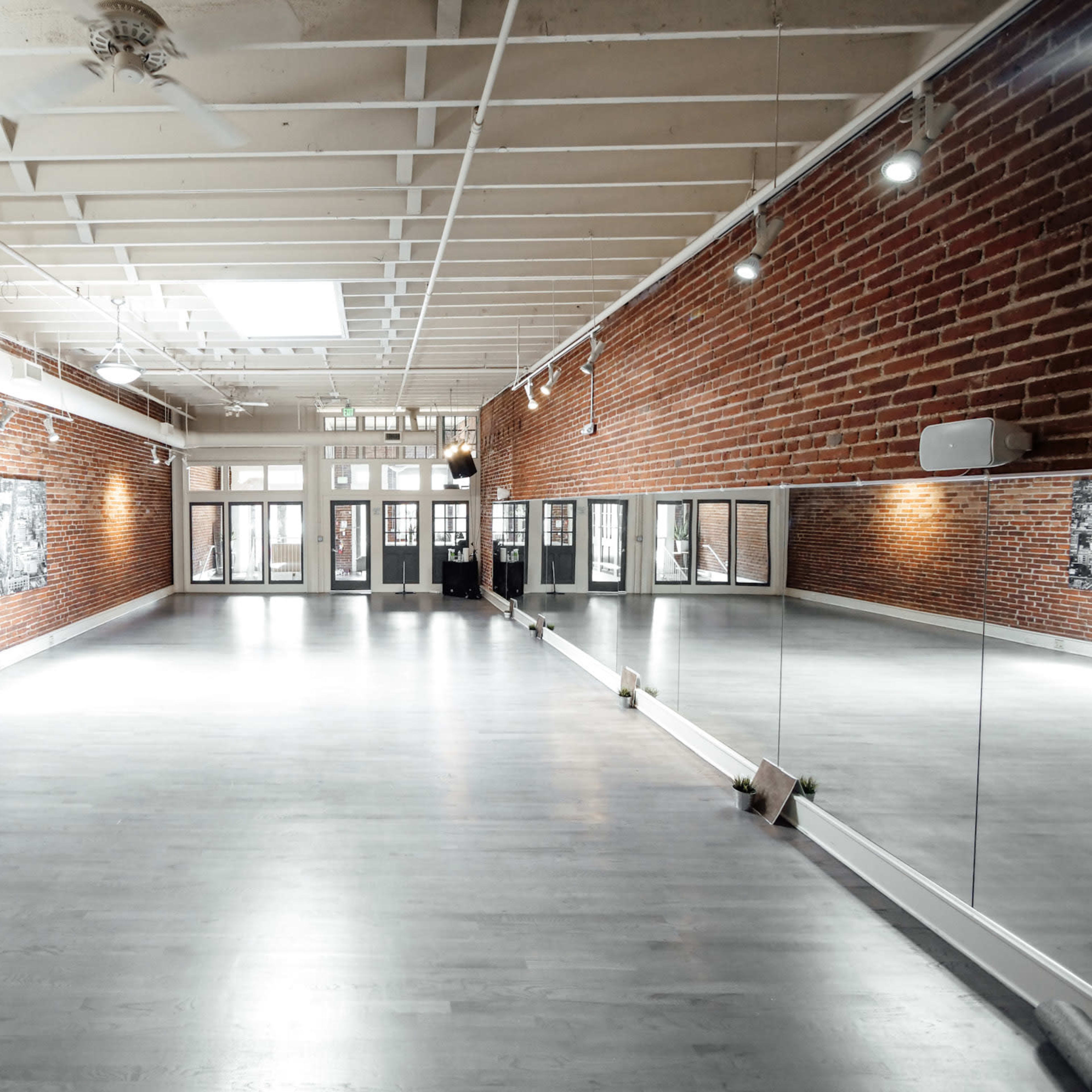 A spacious, well-lit studio with exposed brick walls and a mirrored wall, featuring large windows that provide natural light.