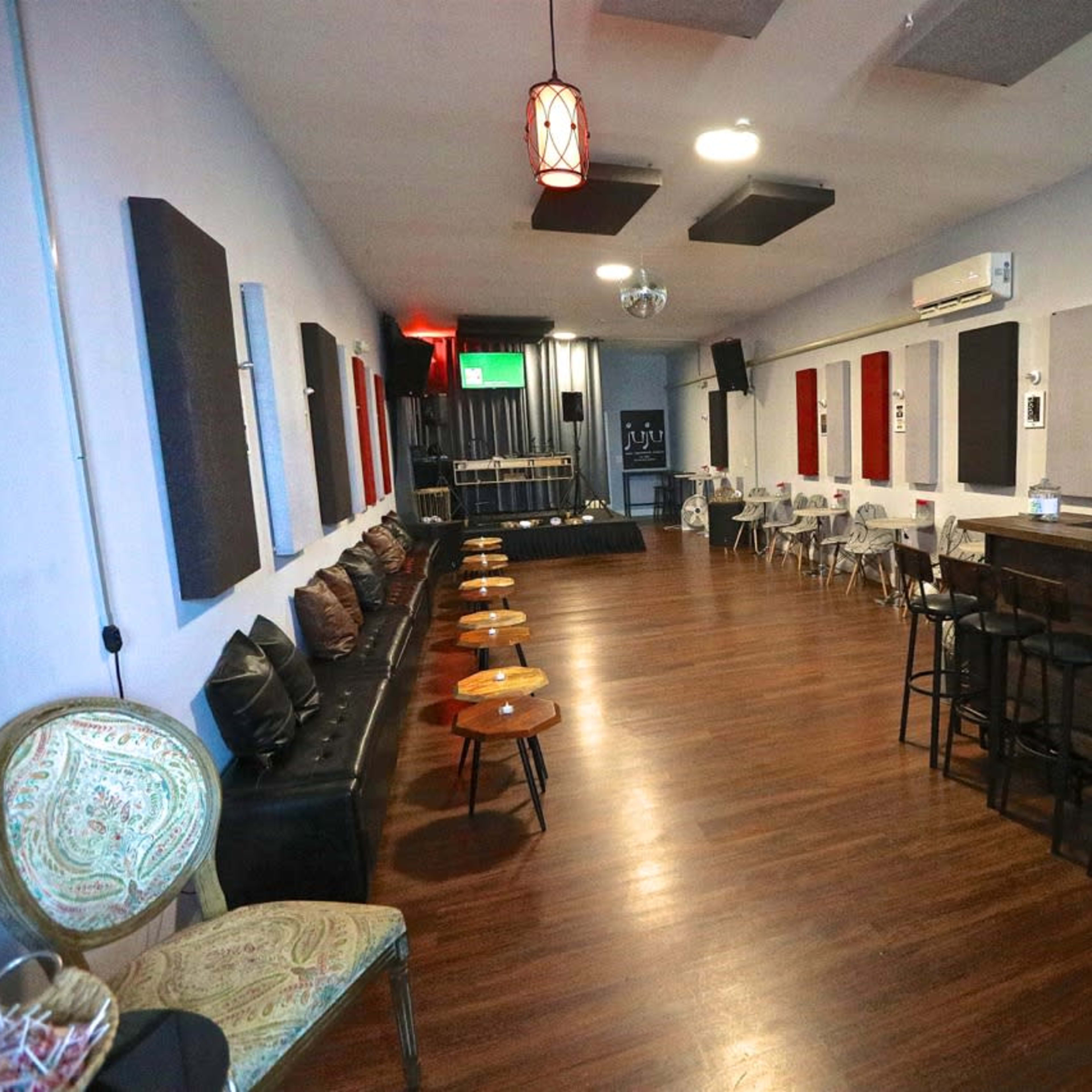 A spacious lounge with a bar, wooden flooring, and chairs arranged around small tables, along with acoustic panels on the walls.