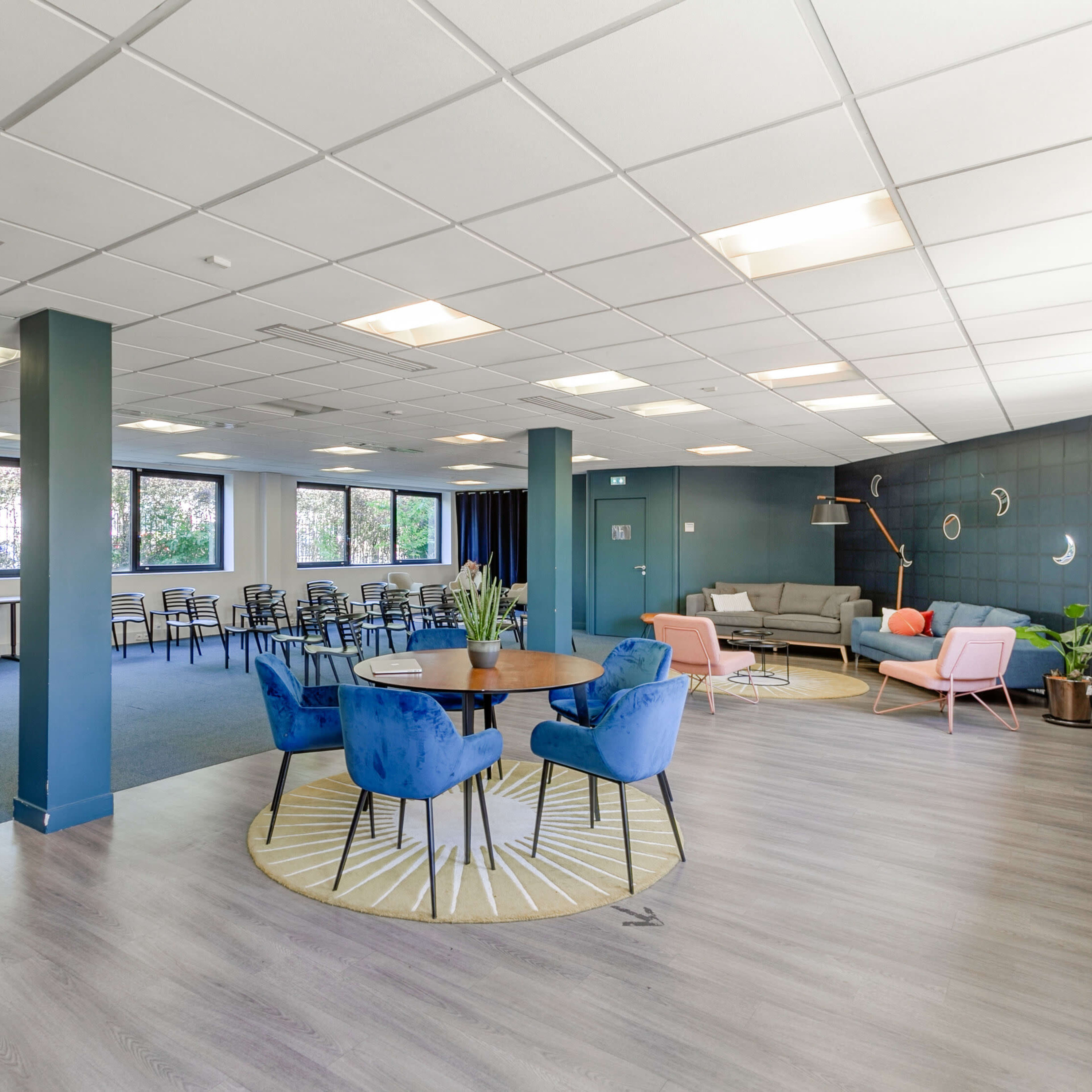 A modern office space with a mix of seating arrangements, including blue chairs and a round table, alongside a lounge area with a sofa and plants.