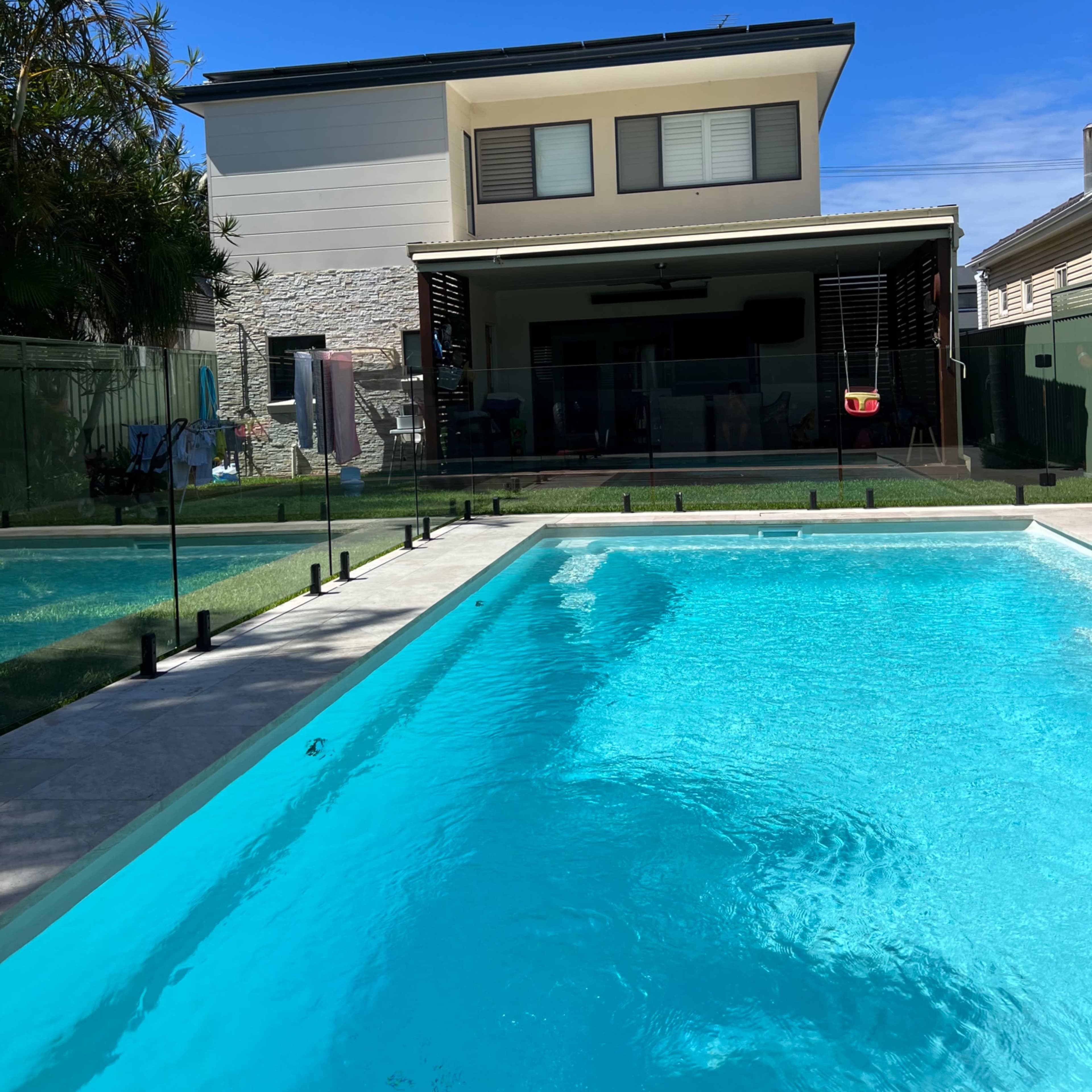 A modern house with a balcony overlooks a clear blue swimming pool surrounded by a grassy yard.