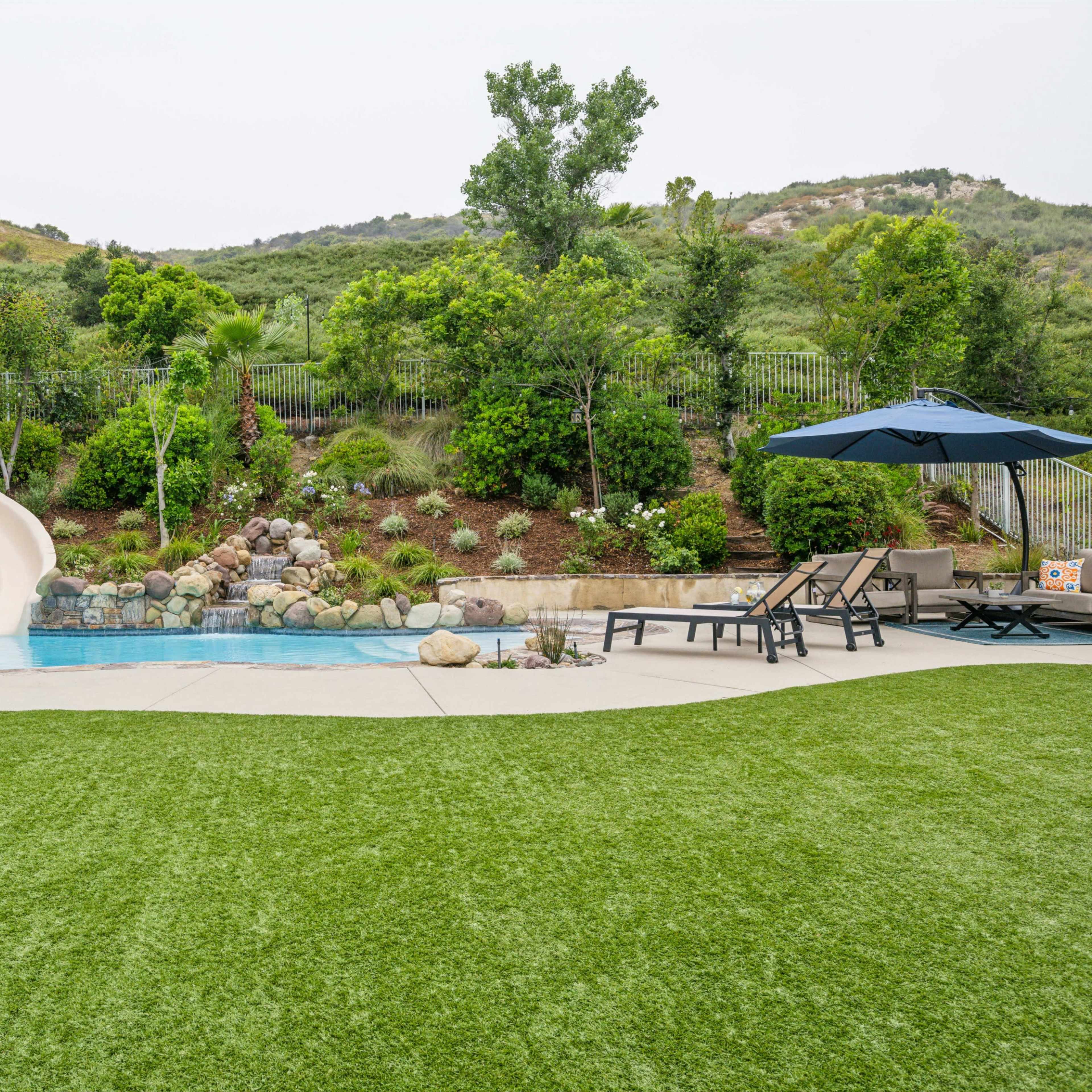 A backyard featuring a swimming pool with a slide, surrounded by landscaped greenery and a seating area under an umbrella.