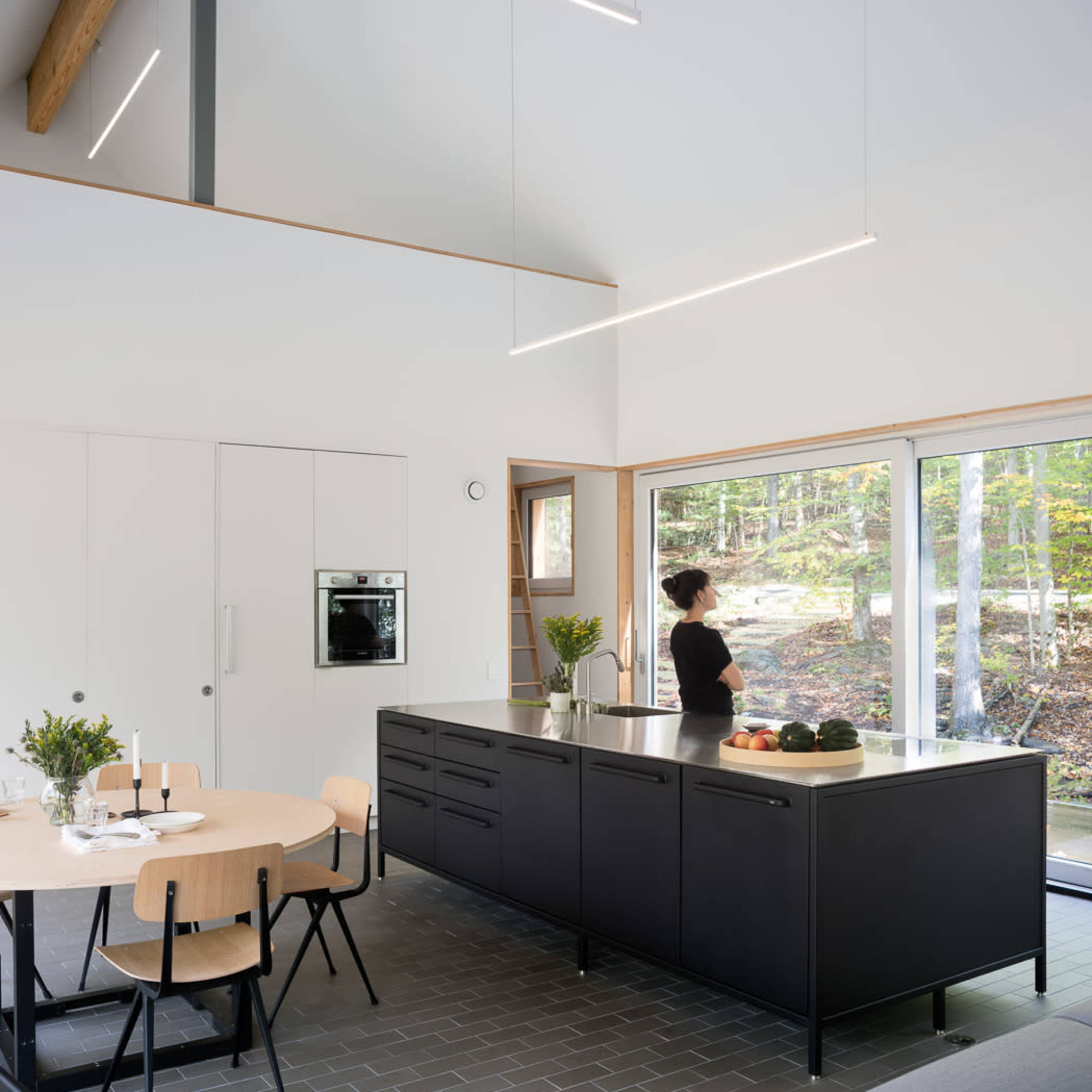 A modern kitchen features a central island with dark cabinetry, a dining table, and large windows that overlook a wooded area.