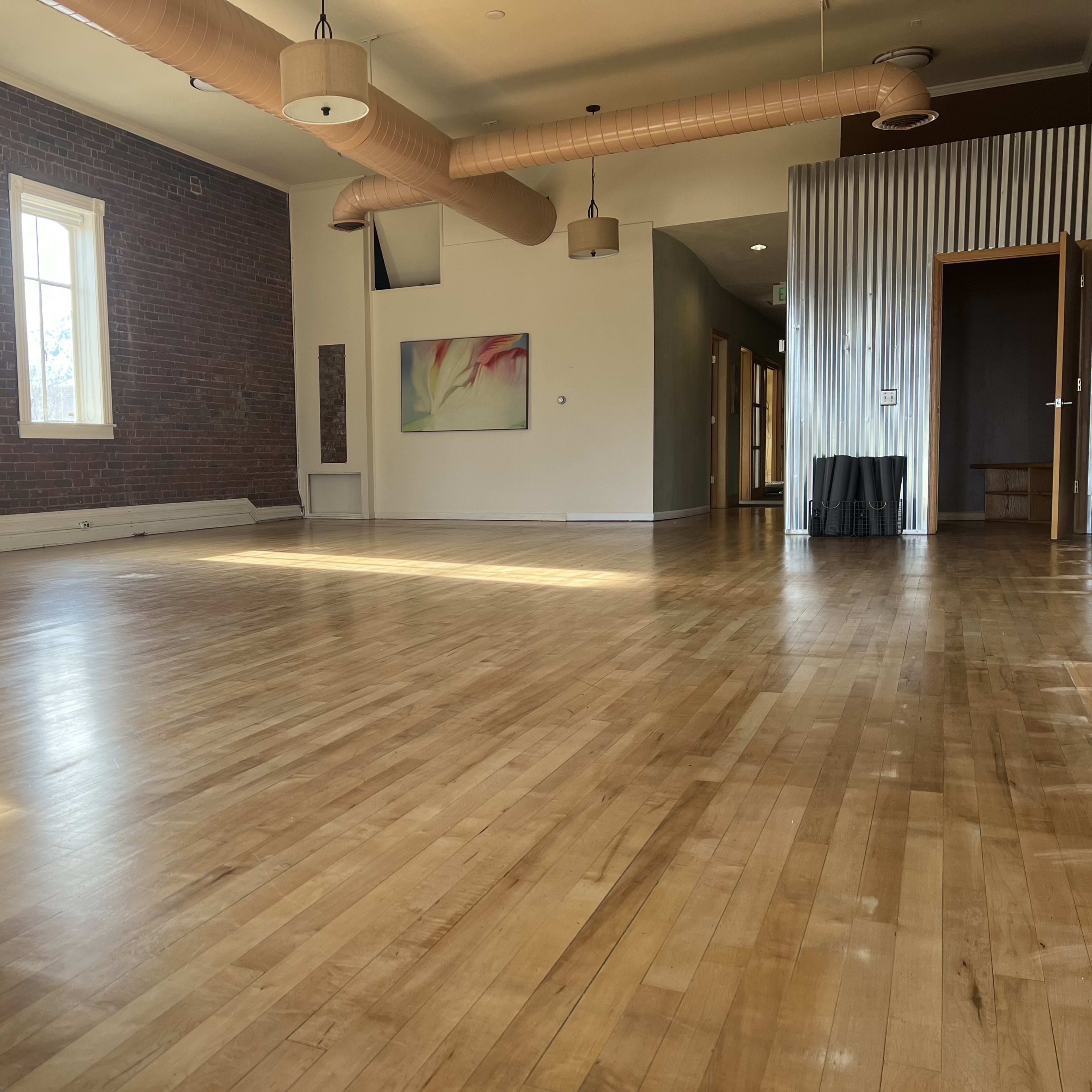 A spacious room with hardwood flooring, large windows, and a few pieces of furniture, including two chairs and a wall art piece.