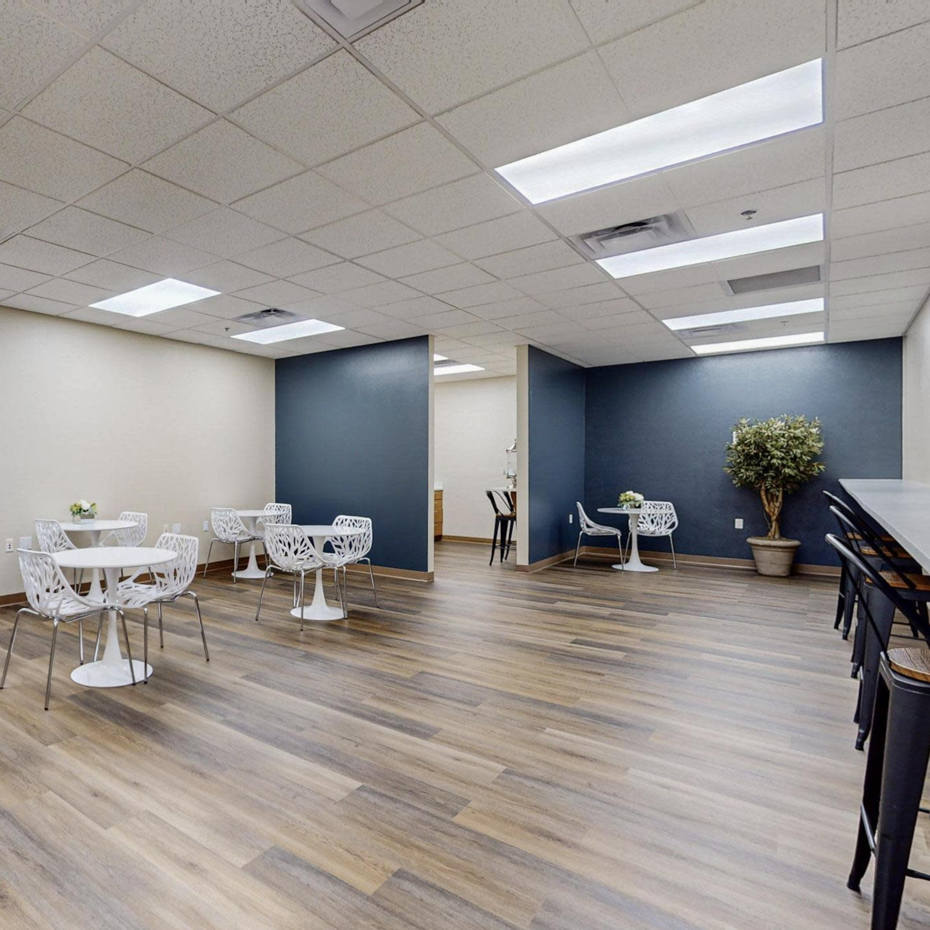 A modern, spacious room with multiple white tables and chairs, a wooden floor, and potted plants along the walls.