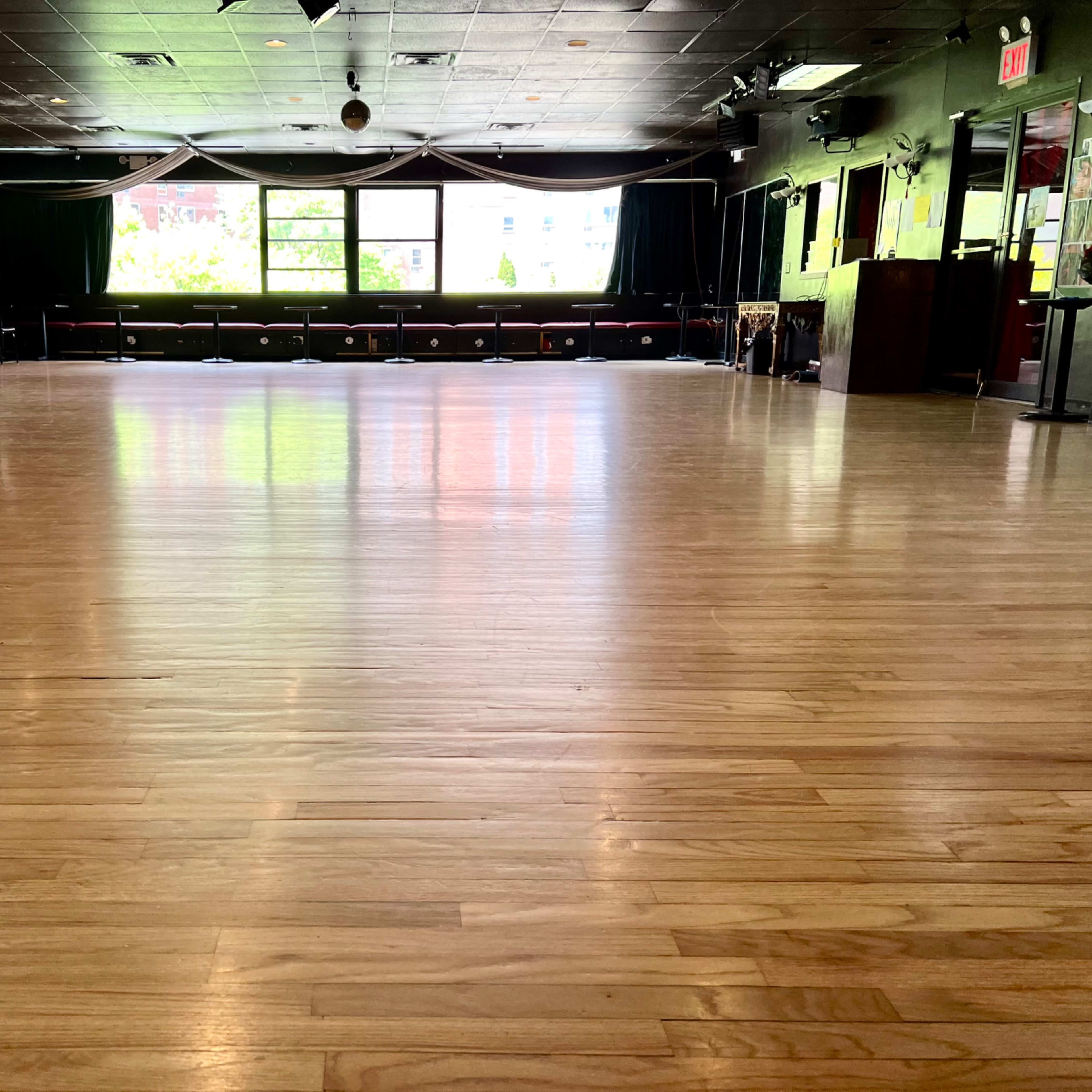 A polished wooden dance floor inside a studio with large windows allowing natural light to enter.