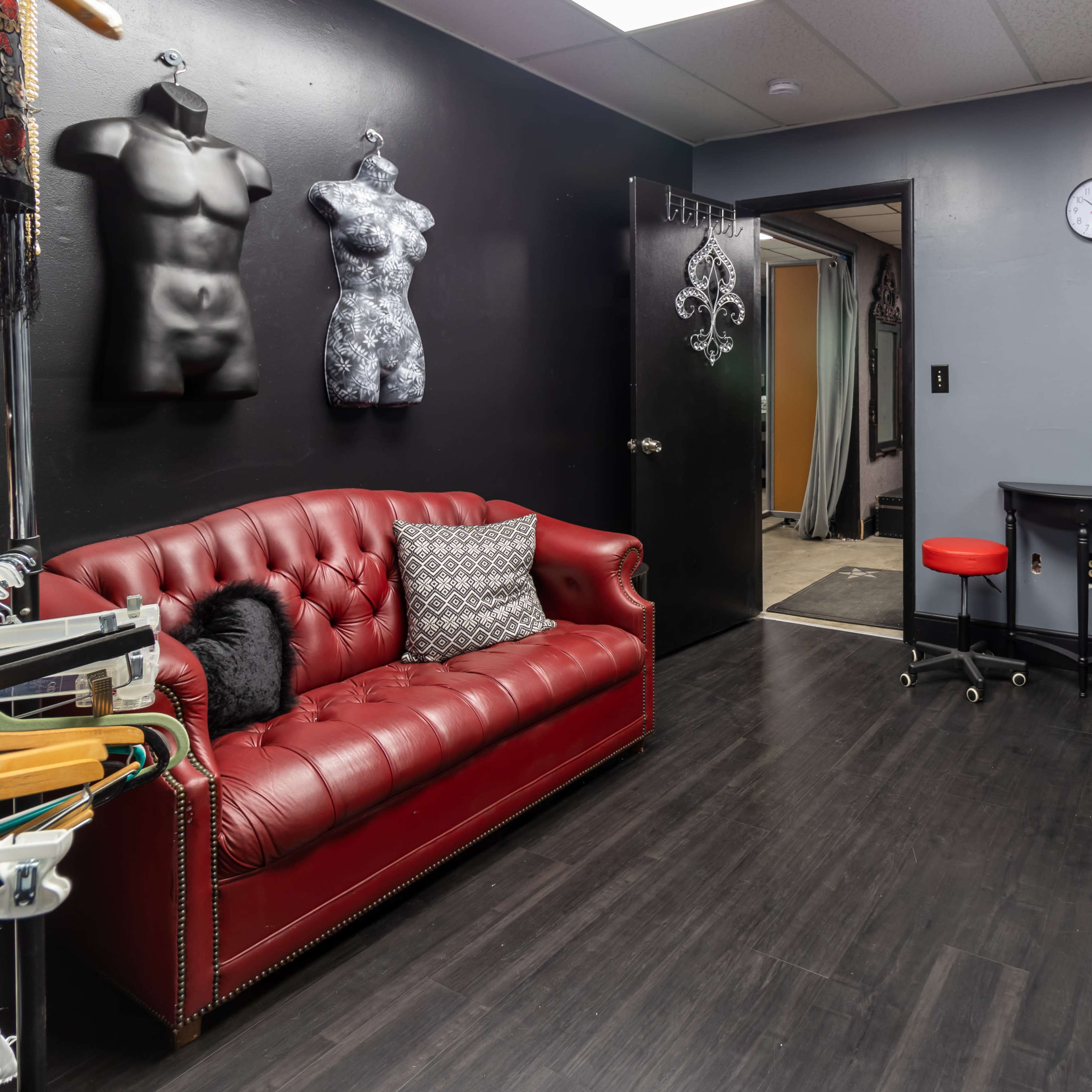 A room with black walls featuring a red leather couch, a small black table, three black barstools, and a mirror with light bulbs, along with mannequins on display.