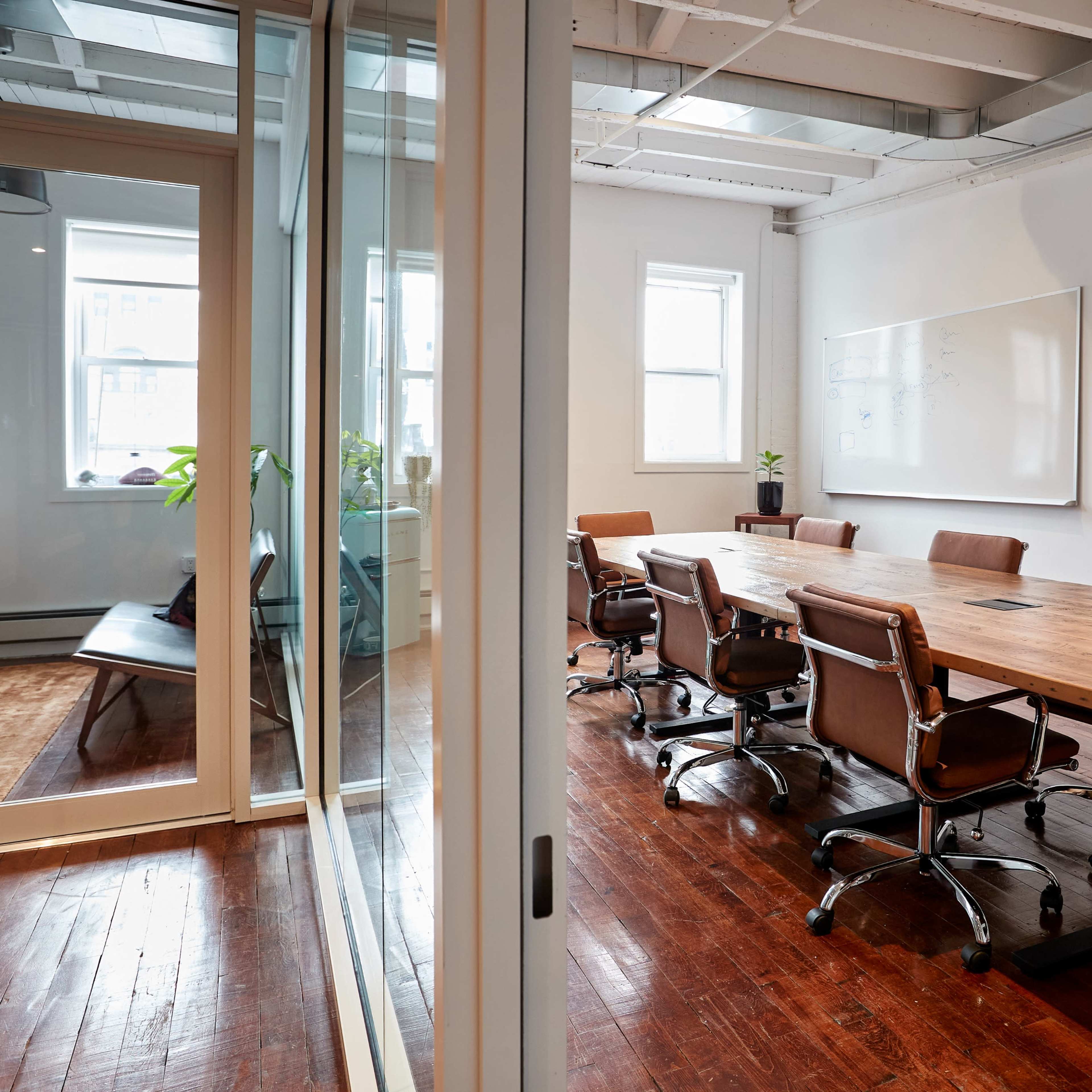 A spacious conference room with a long wooden table surrounded by brown leather swivel chairs, leading to a glass-walled meeting area.