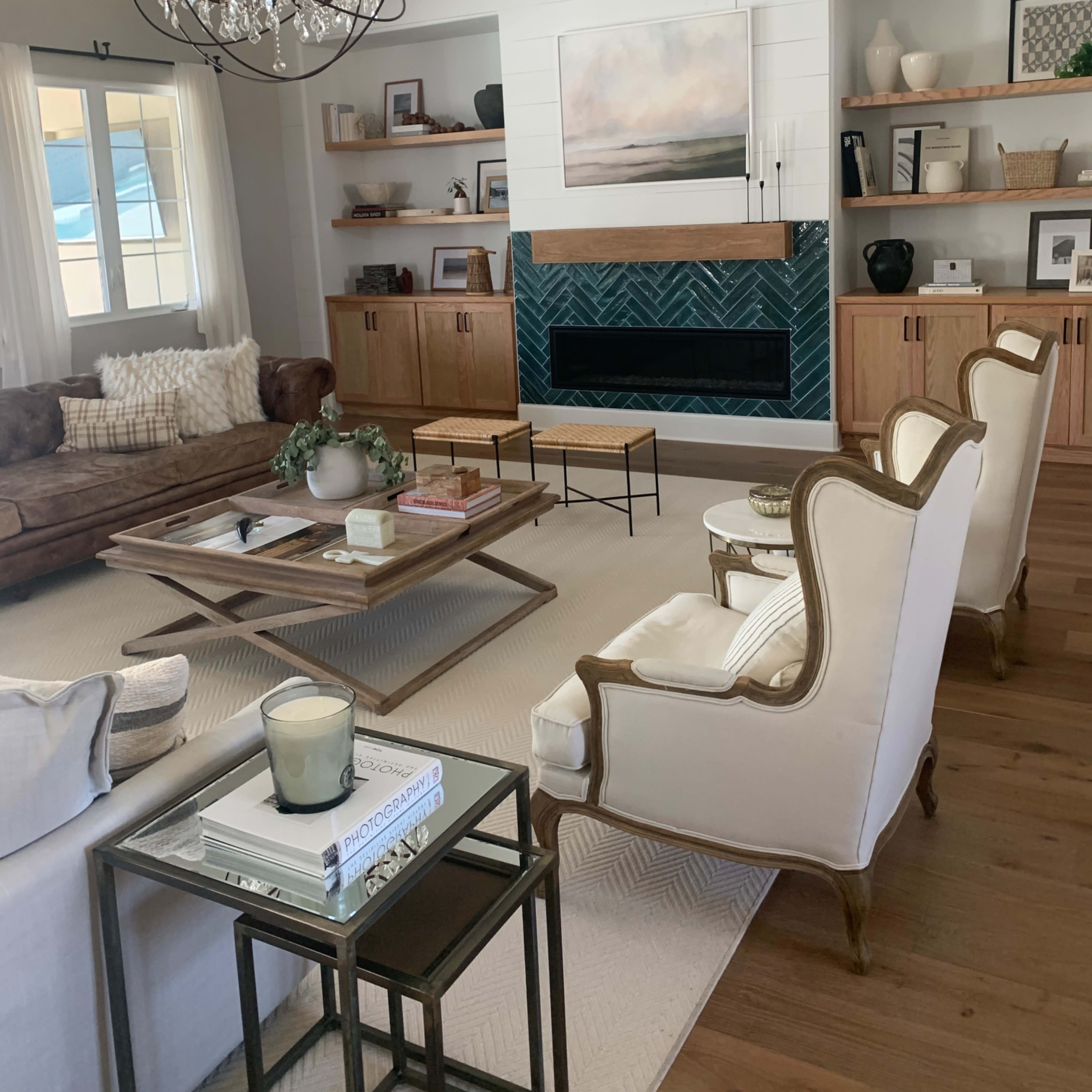The living room features a stylish arrangement with a brown leather sofa, white armchairs, a wooden coffee table, and a modern chandelier above a green tiled fireplace.