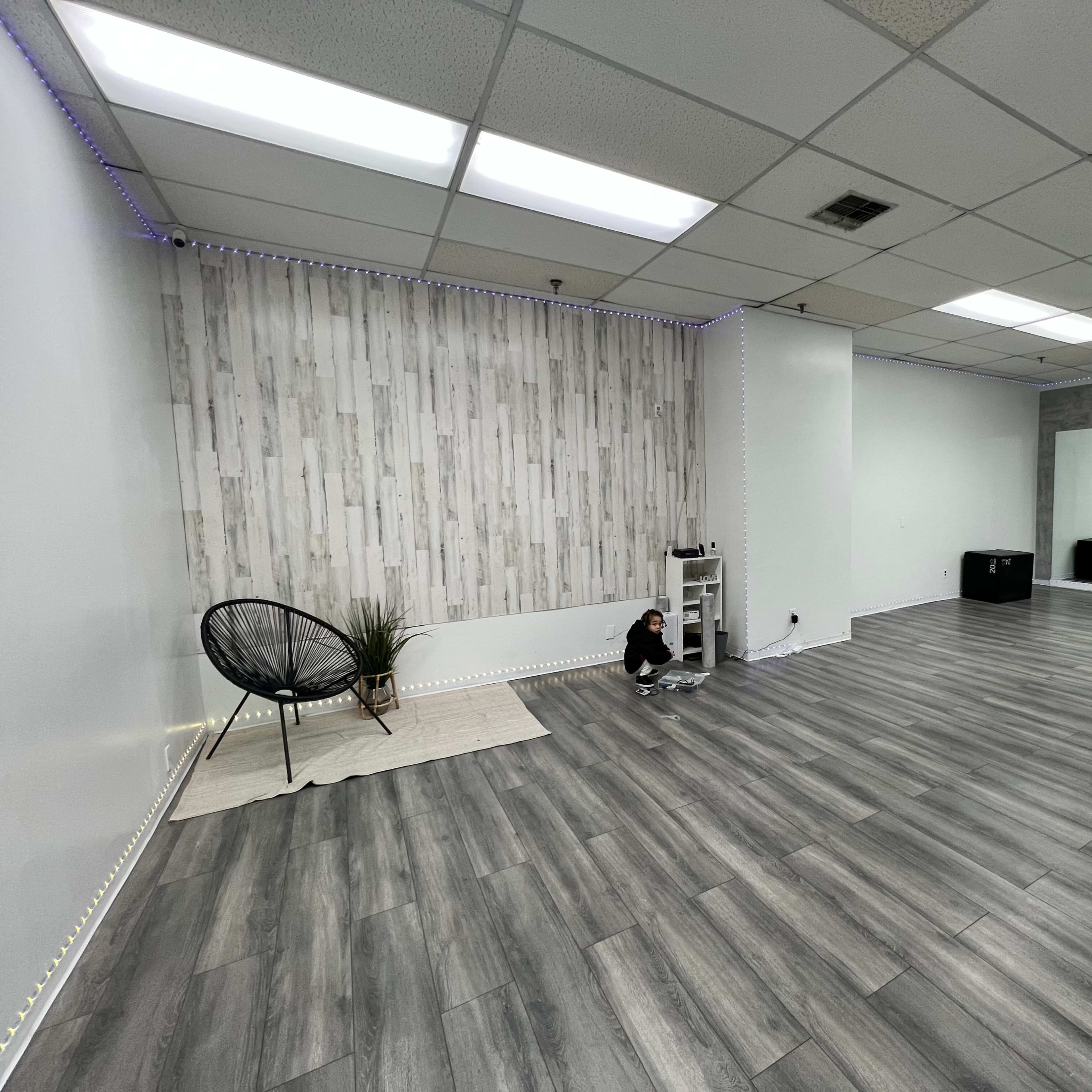 A spacious, empty studio with light gray wood flooring, a decorative wall featuring wood paneling, and a small black chair in a corner.