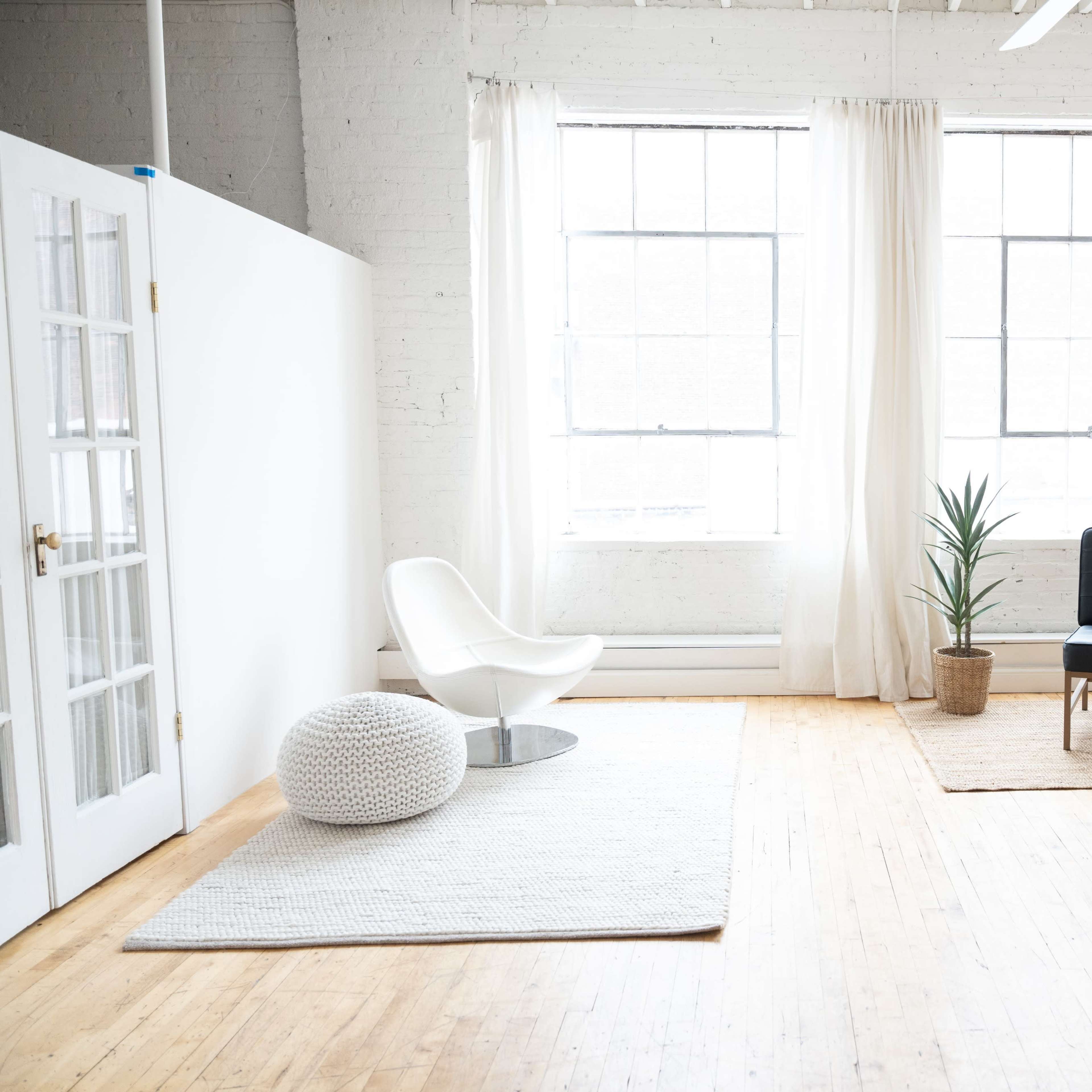A spacious room with large windows, featuring a white chair, a black couch, a potted plant, and rugs on a wooden floor.