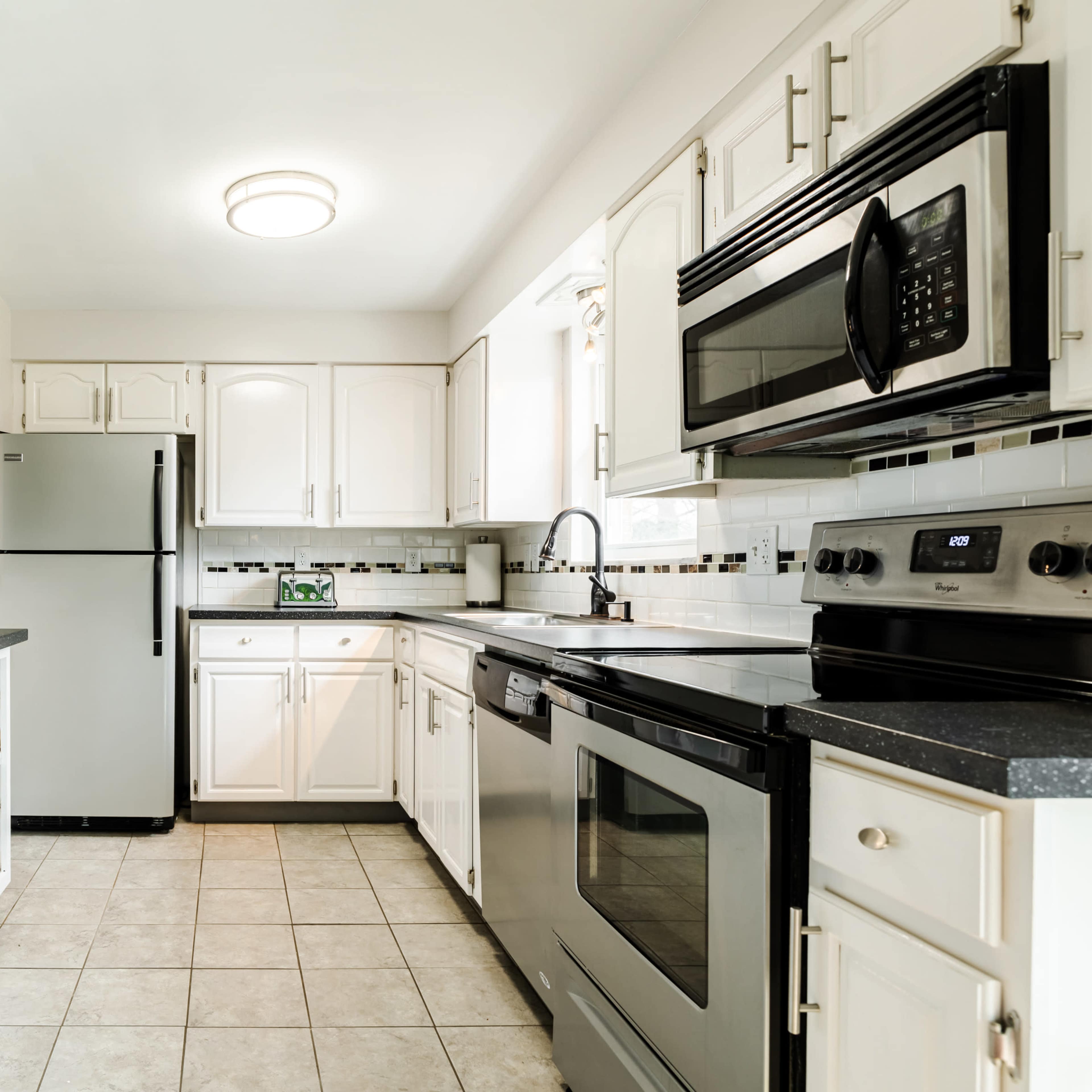 The kitchen features white cabinets, stainless steel appliances, and a tiled floor, with a small shelf displaying a plant and coffee maker.