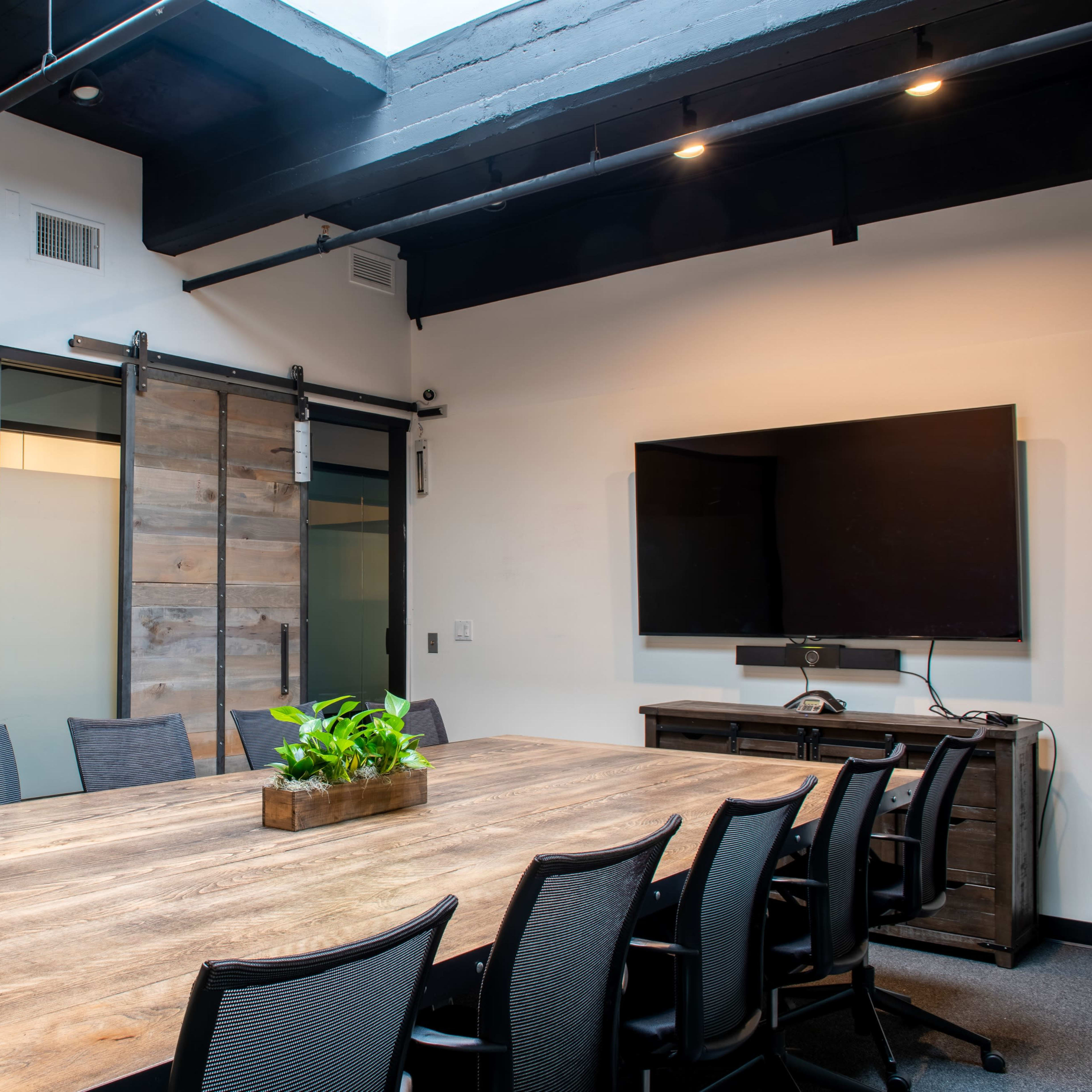 A large wooden conference table surrounded by black chairs is set in a modern meeting room with glass walls and a television on one side.