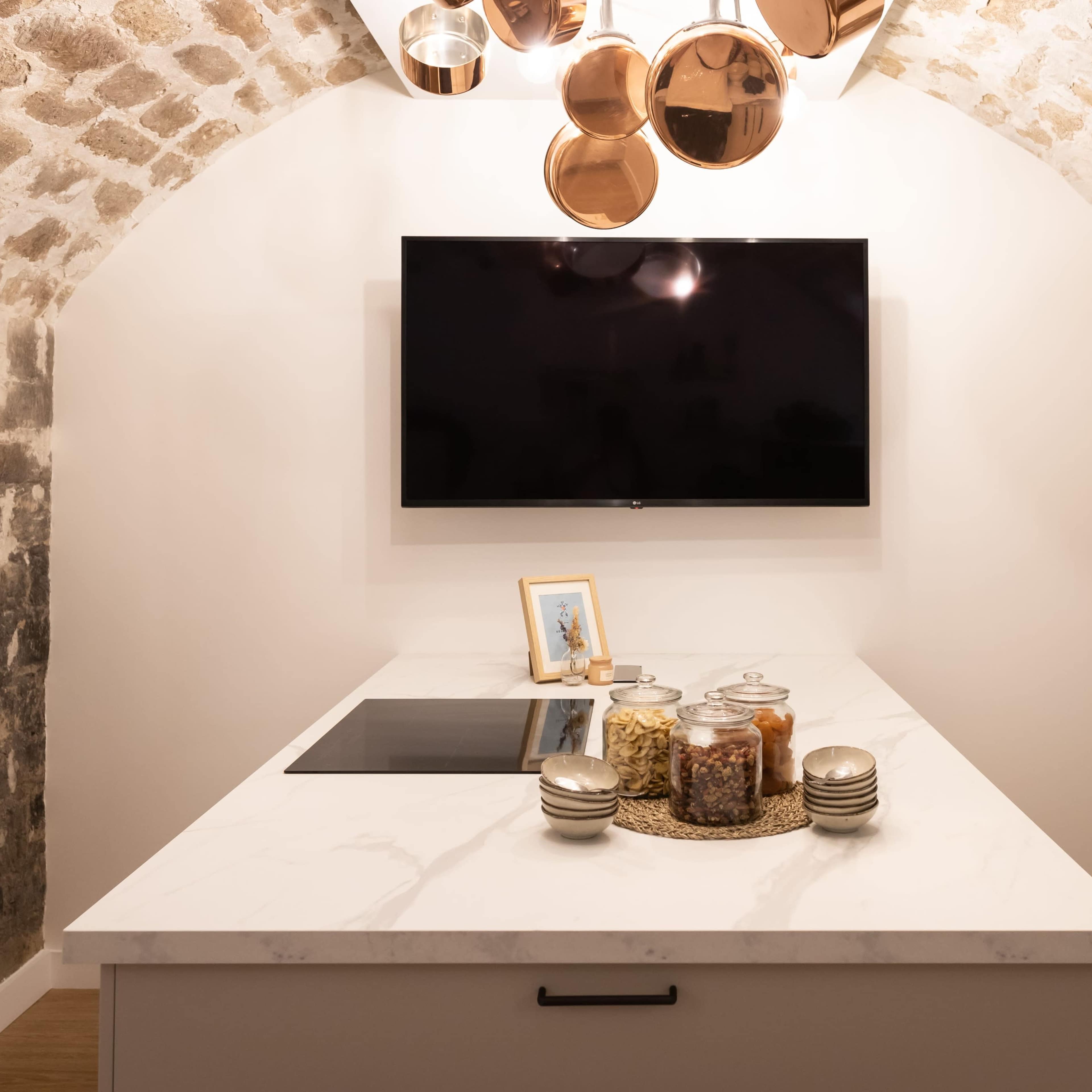 A modern kitchen features a marble island with a built-in cooktop, a wall-mounted television, and copper pendant lights hanging from an arched ceiling.