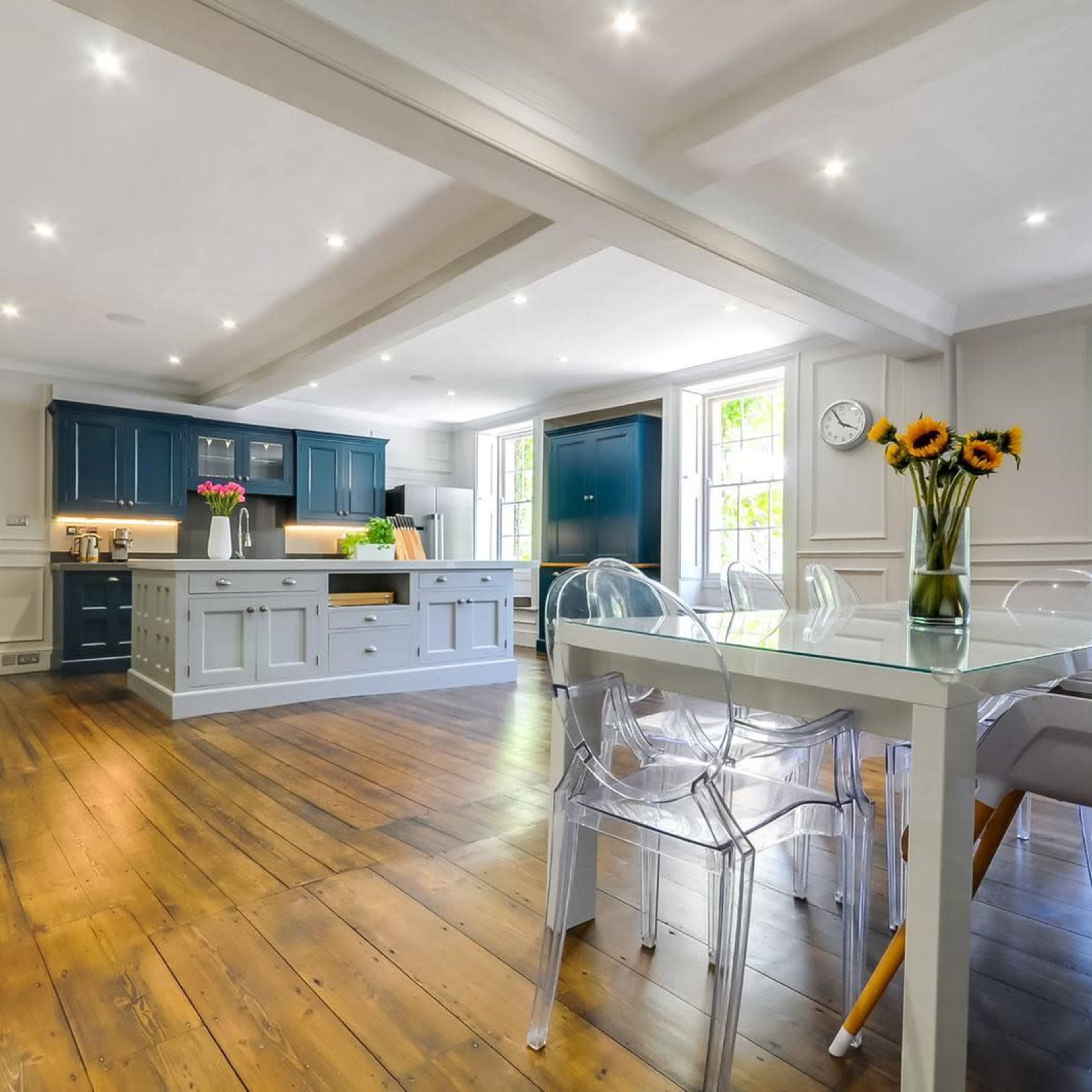A modern kitchen and dining area with wooden flooring, a central island, and a glass dining table surrounded by transparent chairs.