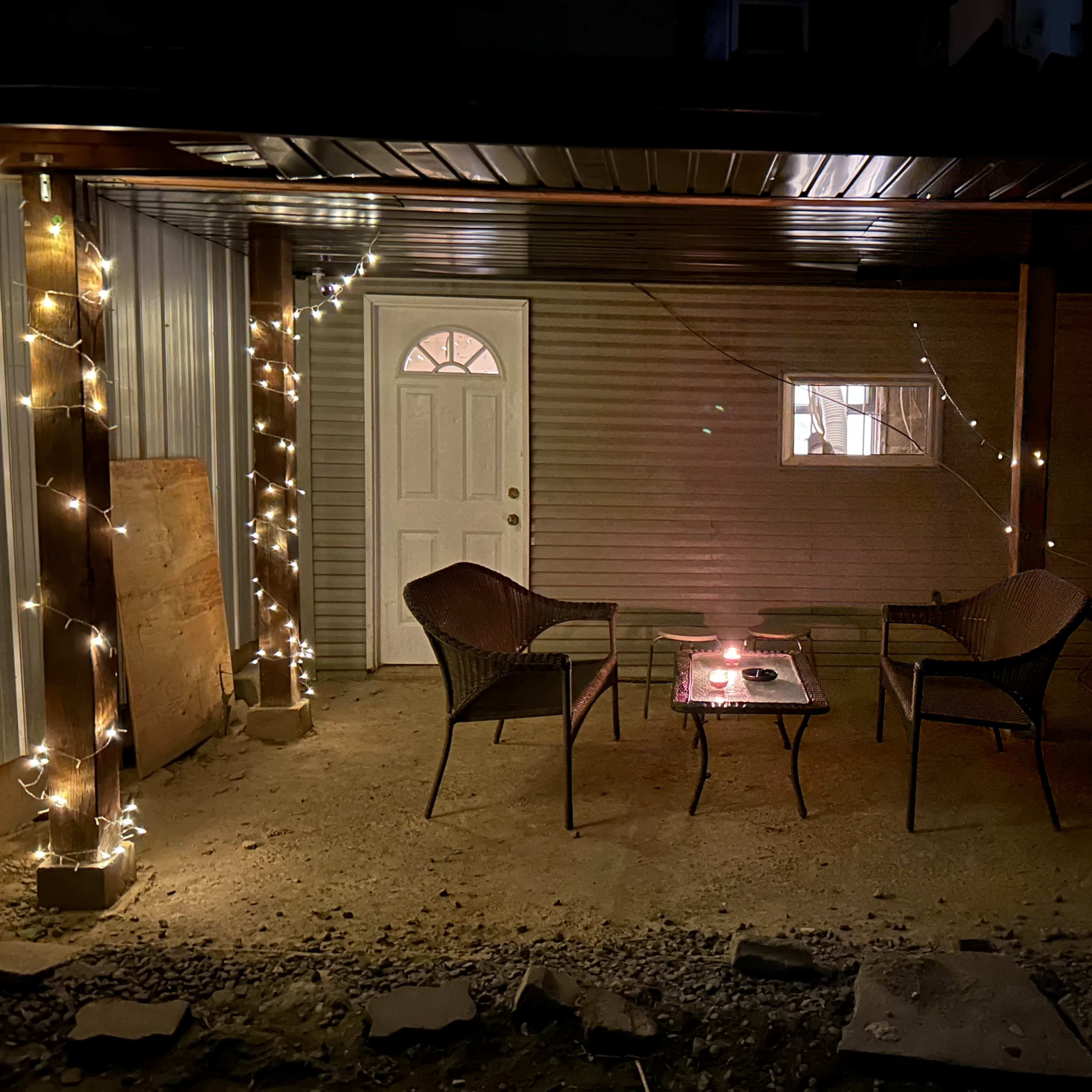 A covered outdoor area features two wicker chairs facing a glass coffee table, adorned with string lights and a door leading to an interior space.