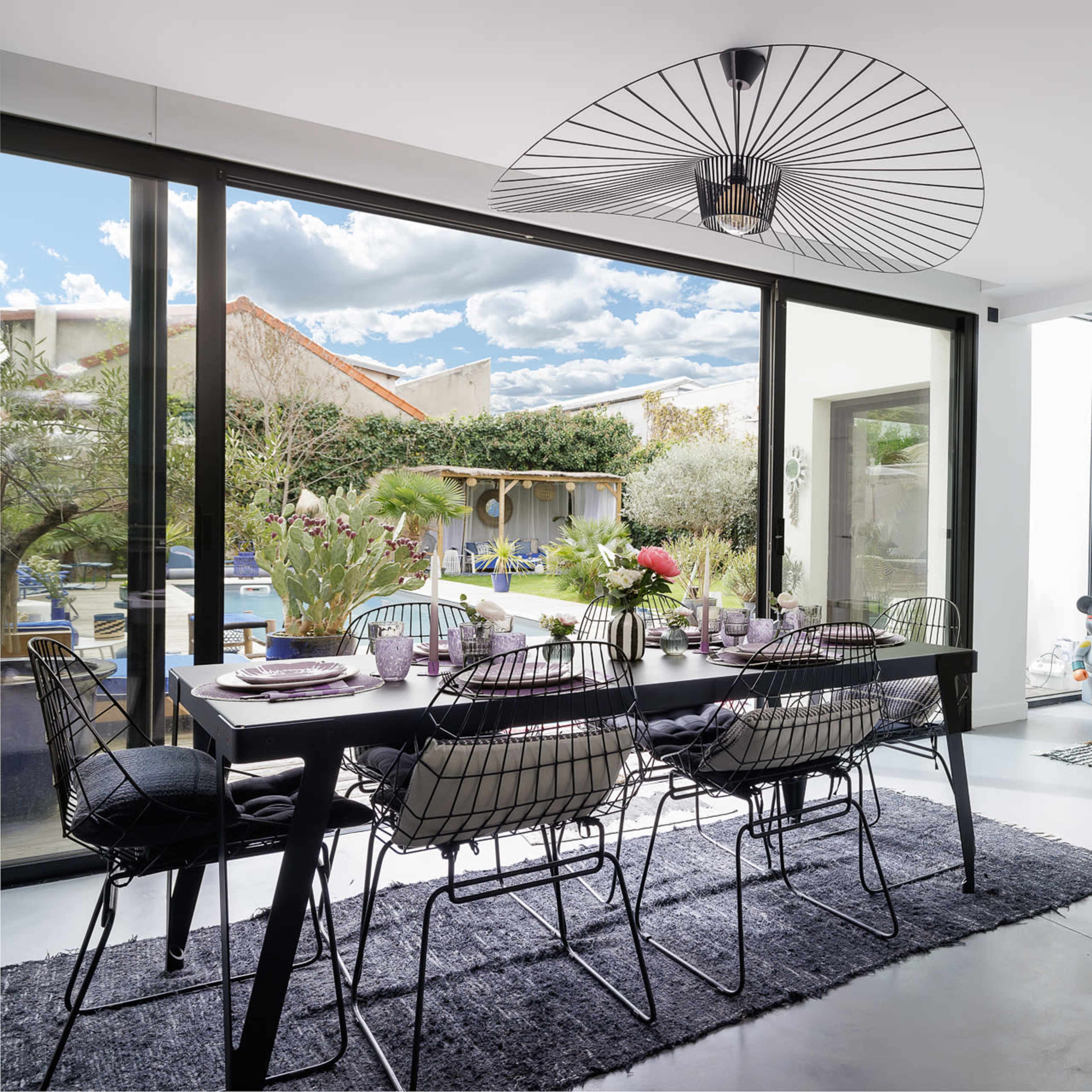 A modern dining area features a black table surrounded by wire chairs, with large windows overlooking a garden and pool.