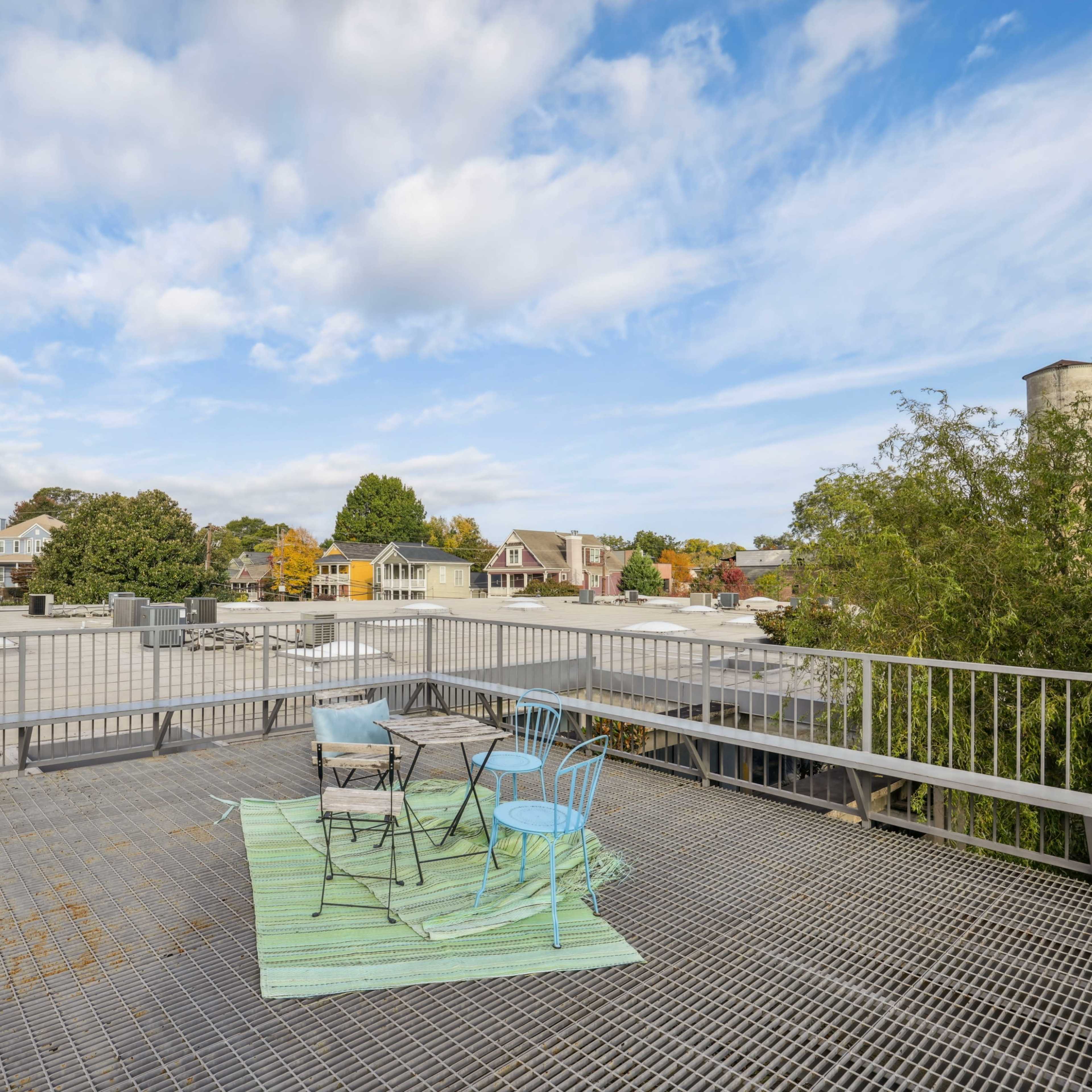 A rooftop terrace with a green rug and four chairs overlooking a neighborhood of houses under a partly cloudy sky.
