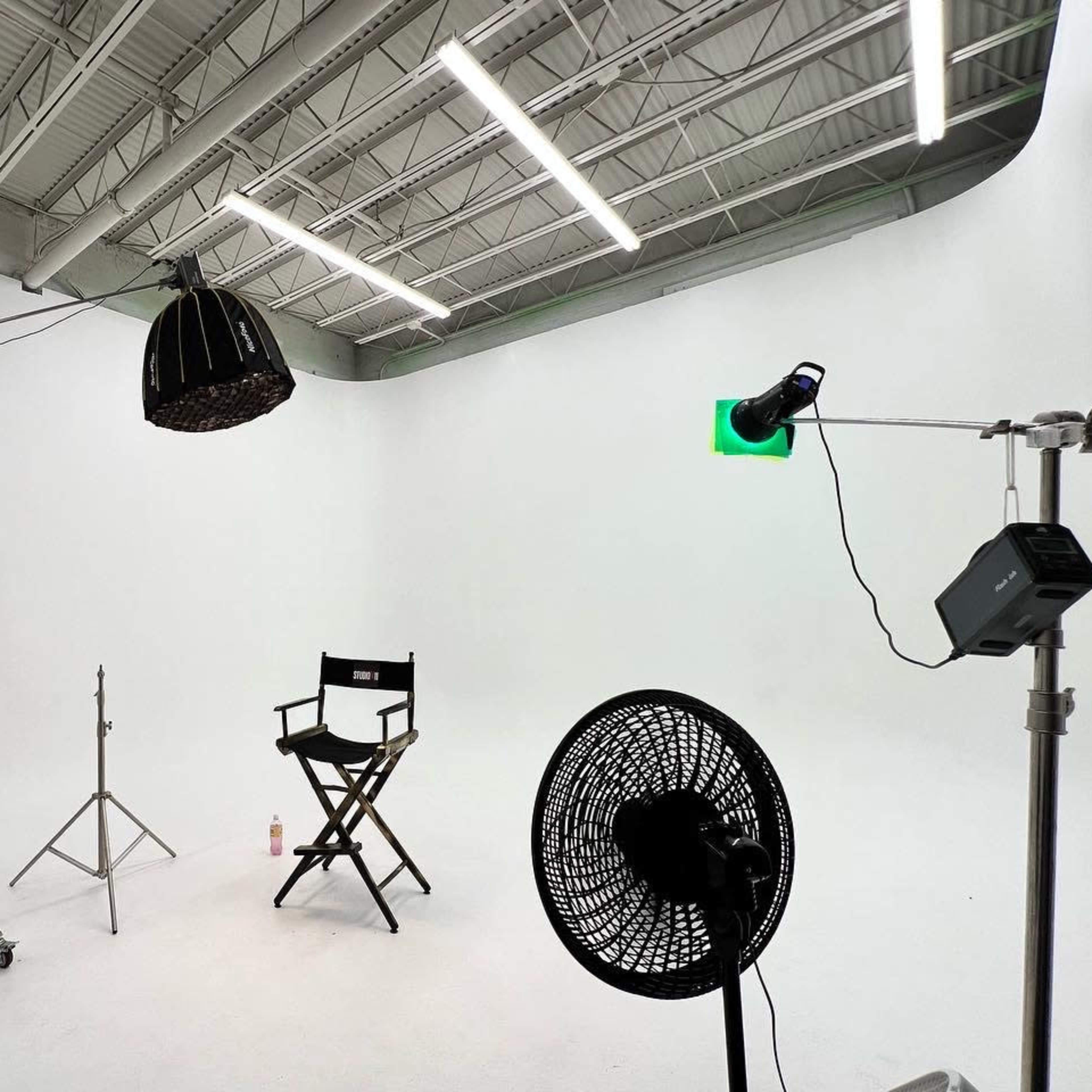 A minimalist photography studio setup with a director's chair, lighting equipment, and a cooling fan against a white background.