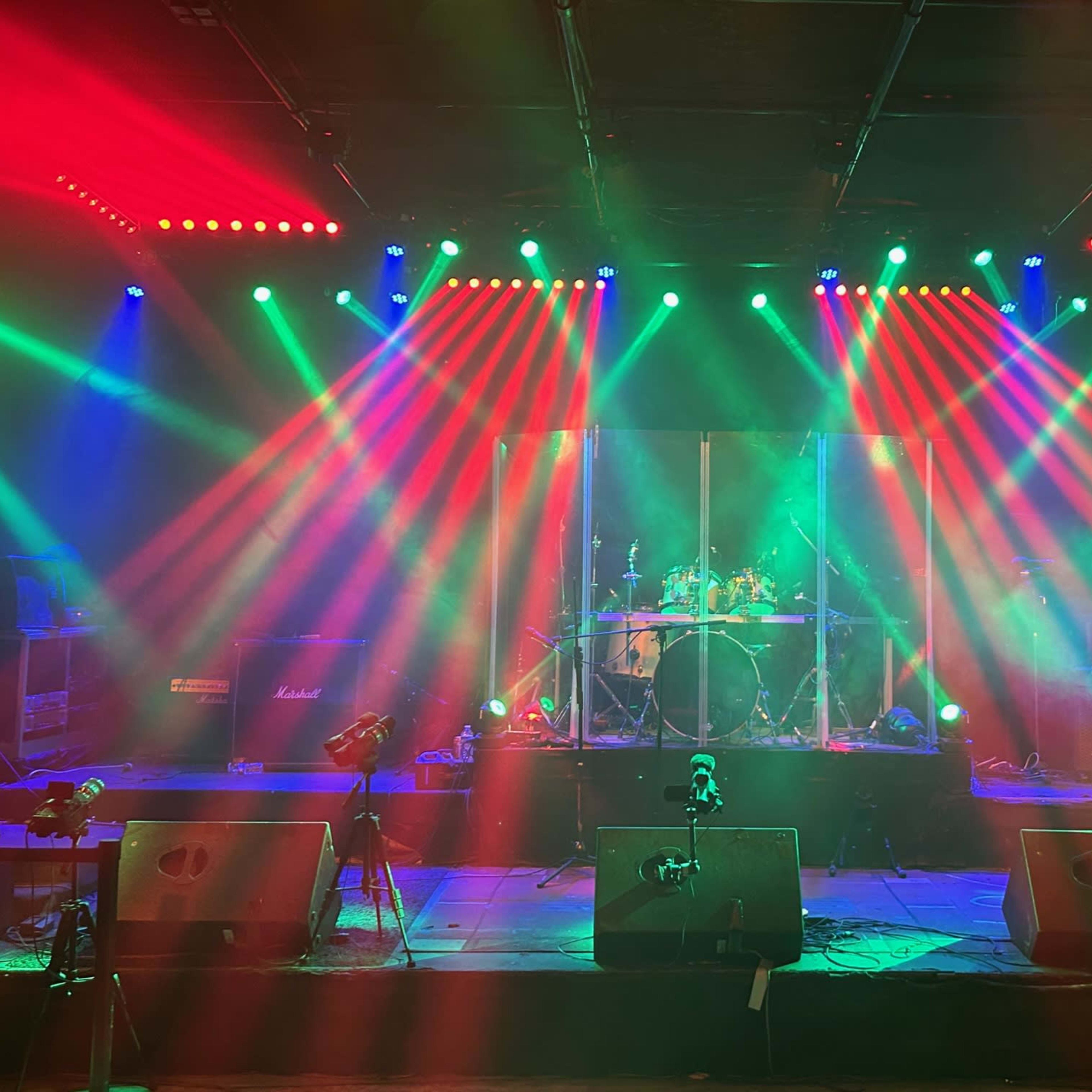 The stage is set with colorful lights shining down on a drum kit, surrounded by amplifiers and microphone stands.