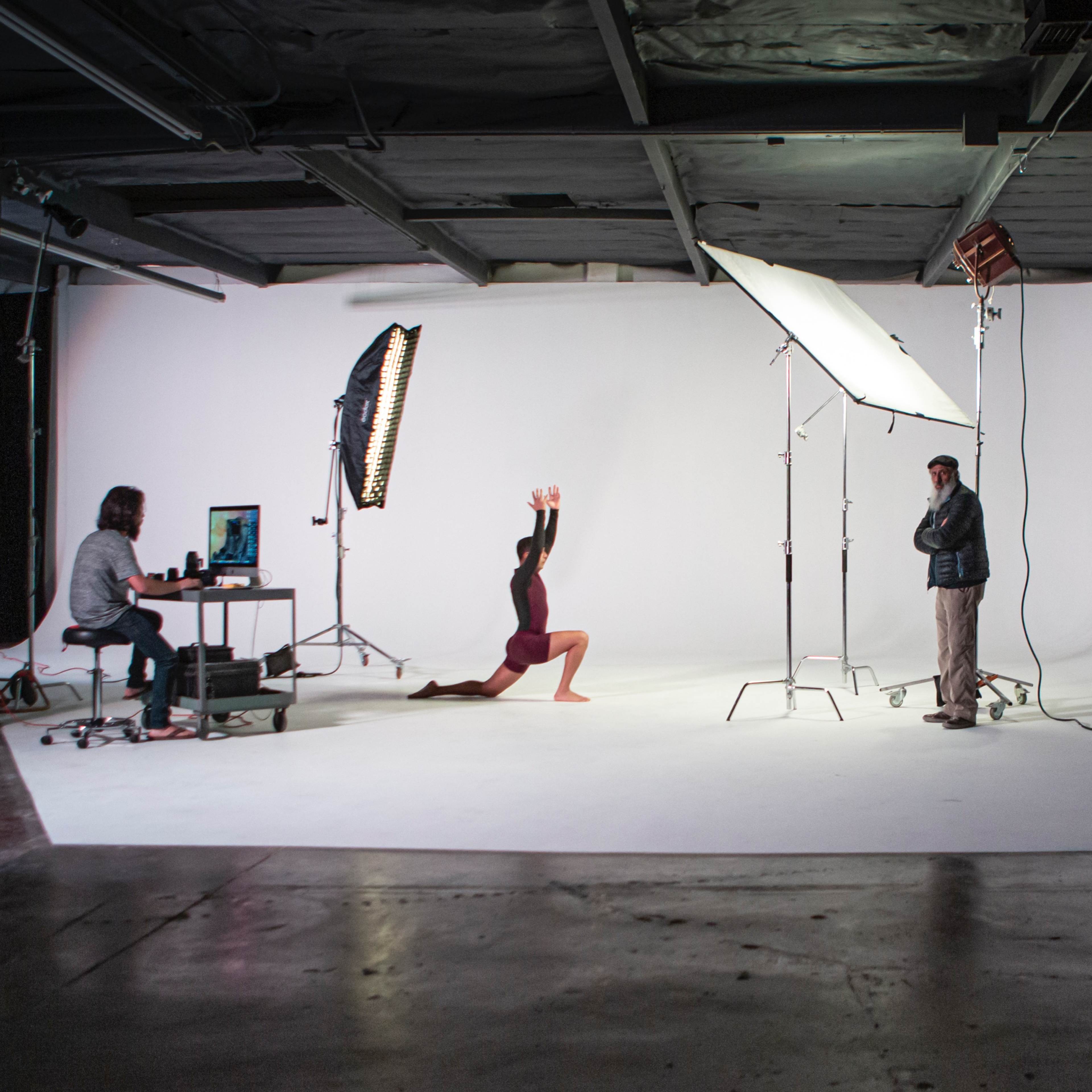 A photoshoot setup features a model posing on her knees in front of a white backdrop, while a photographer and an assistant adjust lighting equipment, and another person operates a camera.