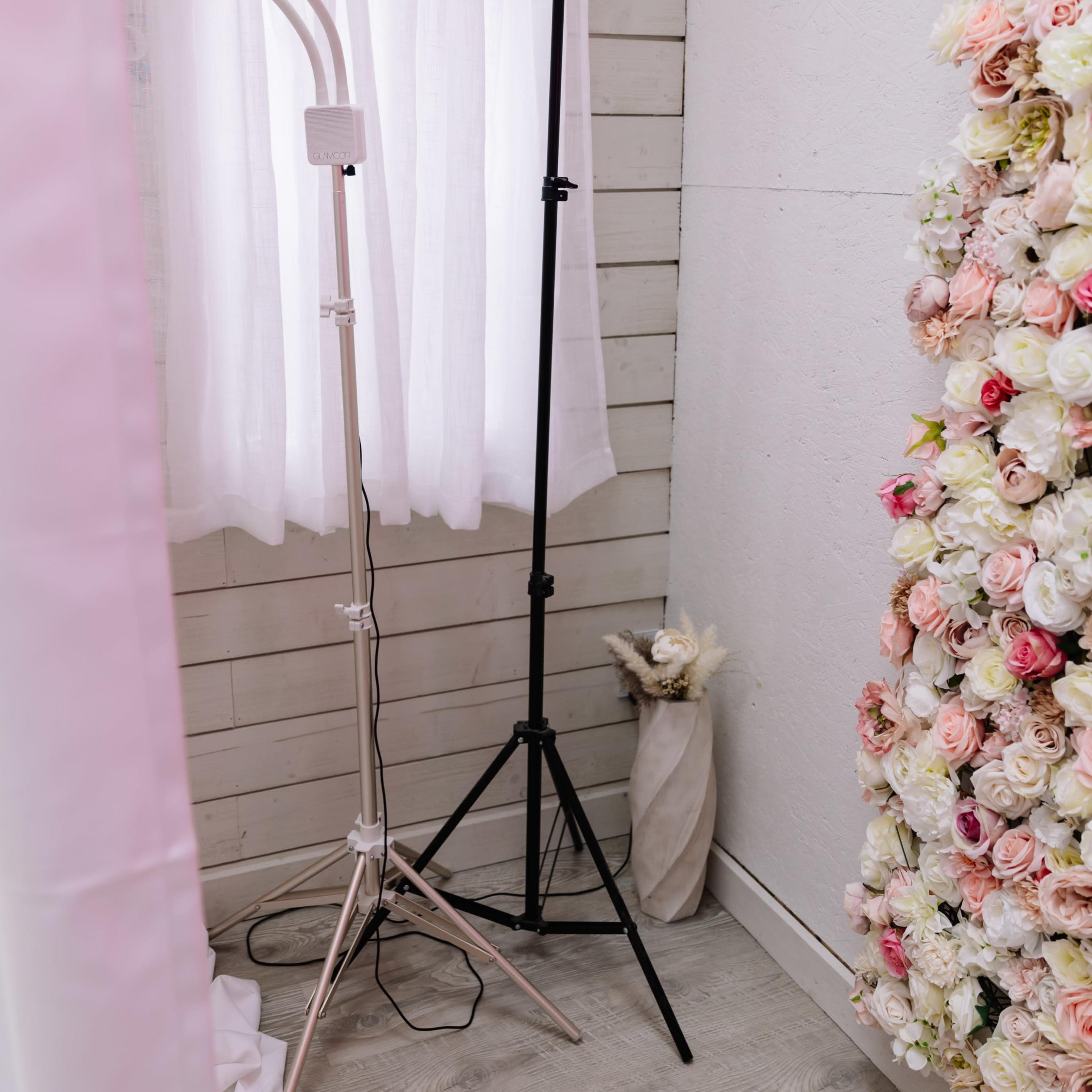 A corner of a room features a black lighting stand next to a tripod light, with white curtains and a wall adorned with flowers.