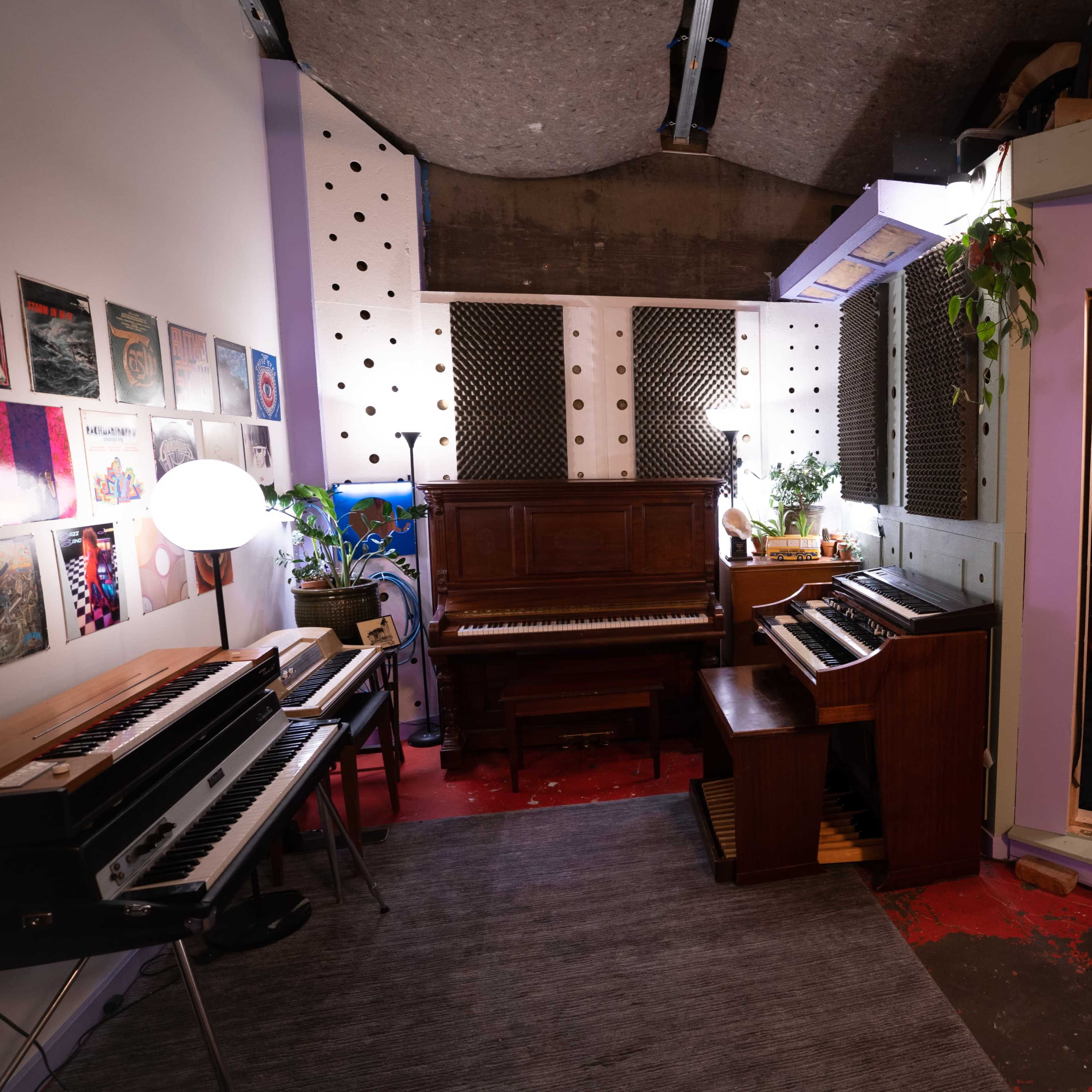 A music studio featuring multiple keyboards, a piano, and a booth for recording, with plants and wall art enhancing the space.