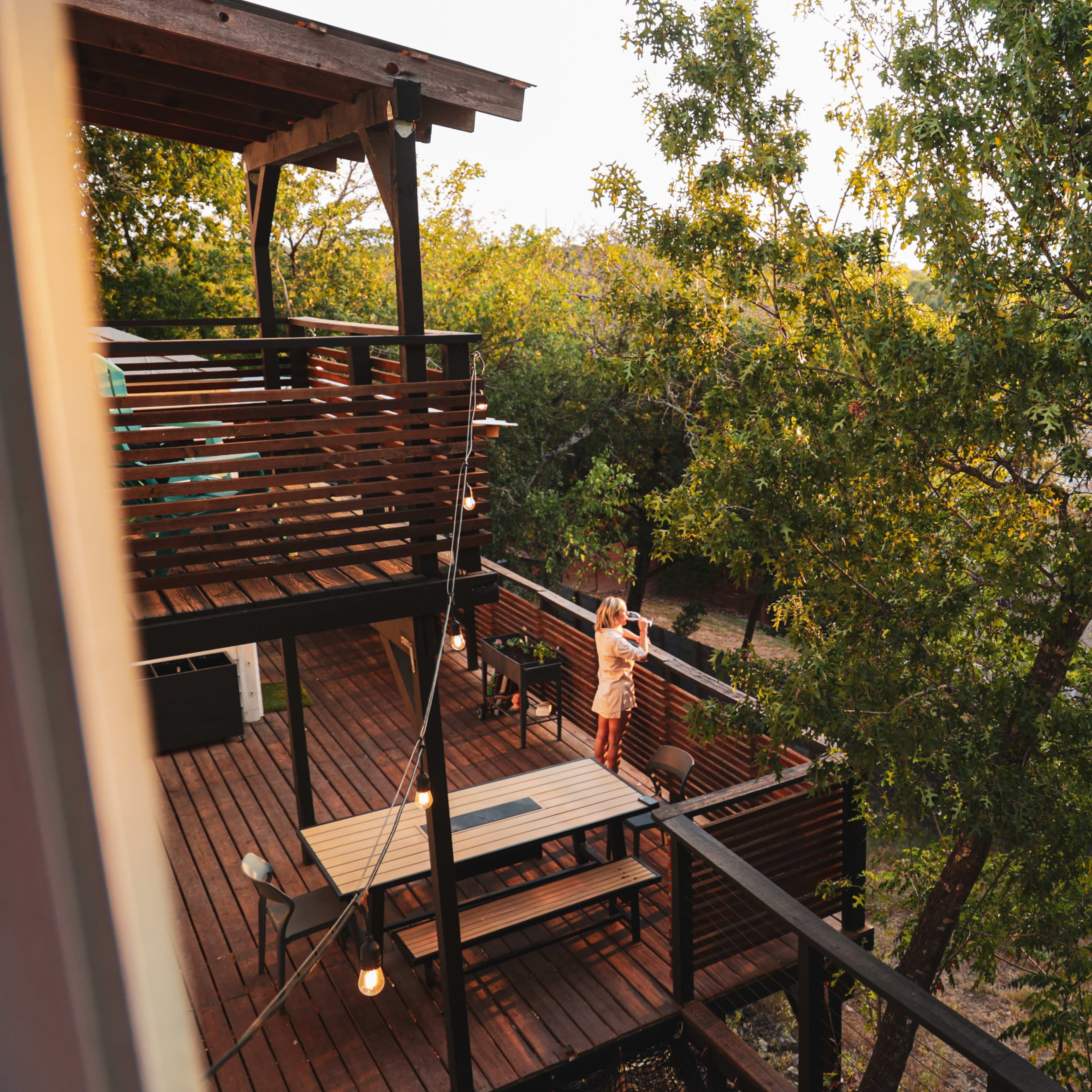 A wooden deck with outdoor seating is surrounded by trees in a residential area during golden hour.