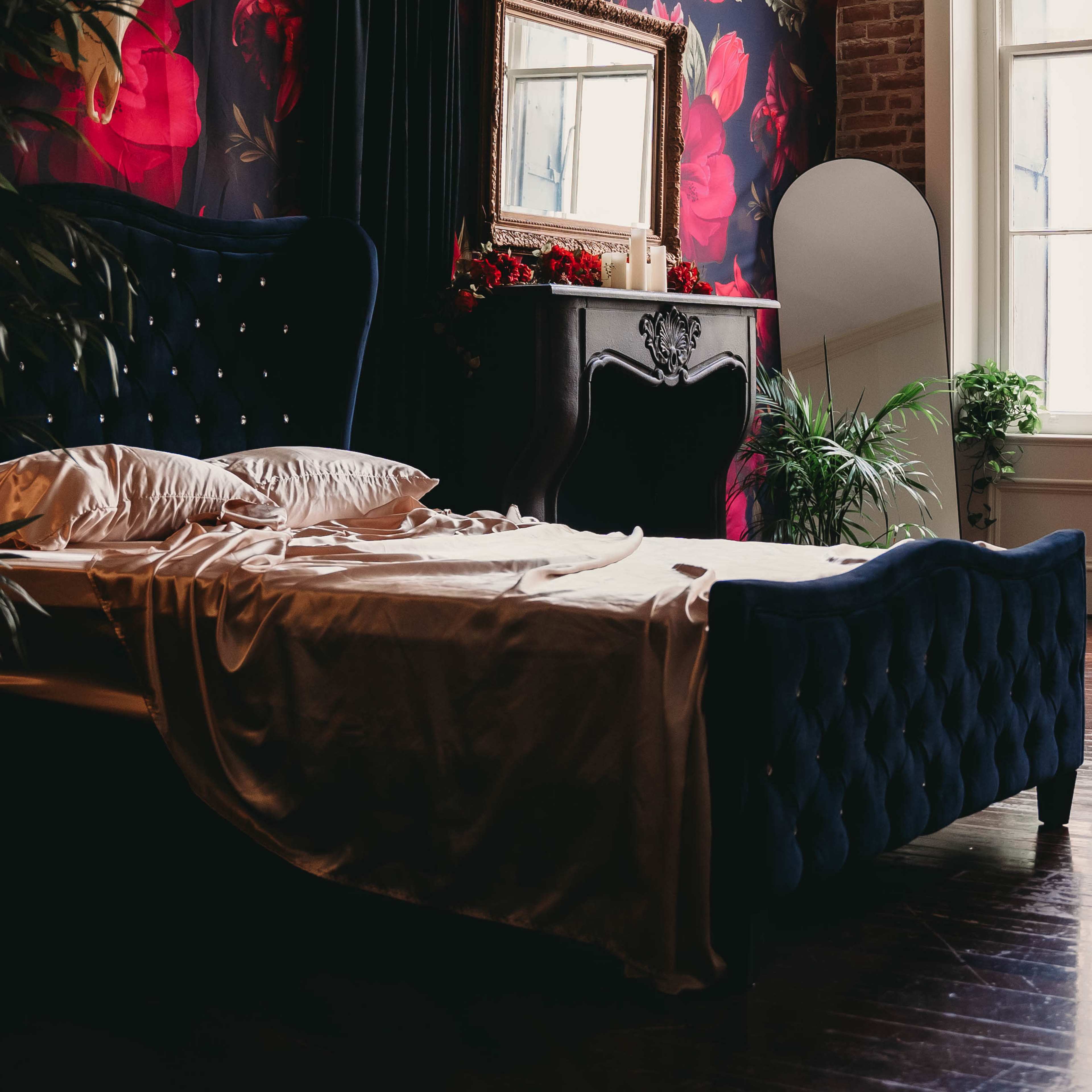 A dark blue upholstered bed sits against a floral wallpaper backdrop, featuring pillows and a crumpled satin blanket, with a vintage mirror and decorative plants nearby.