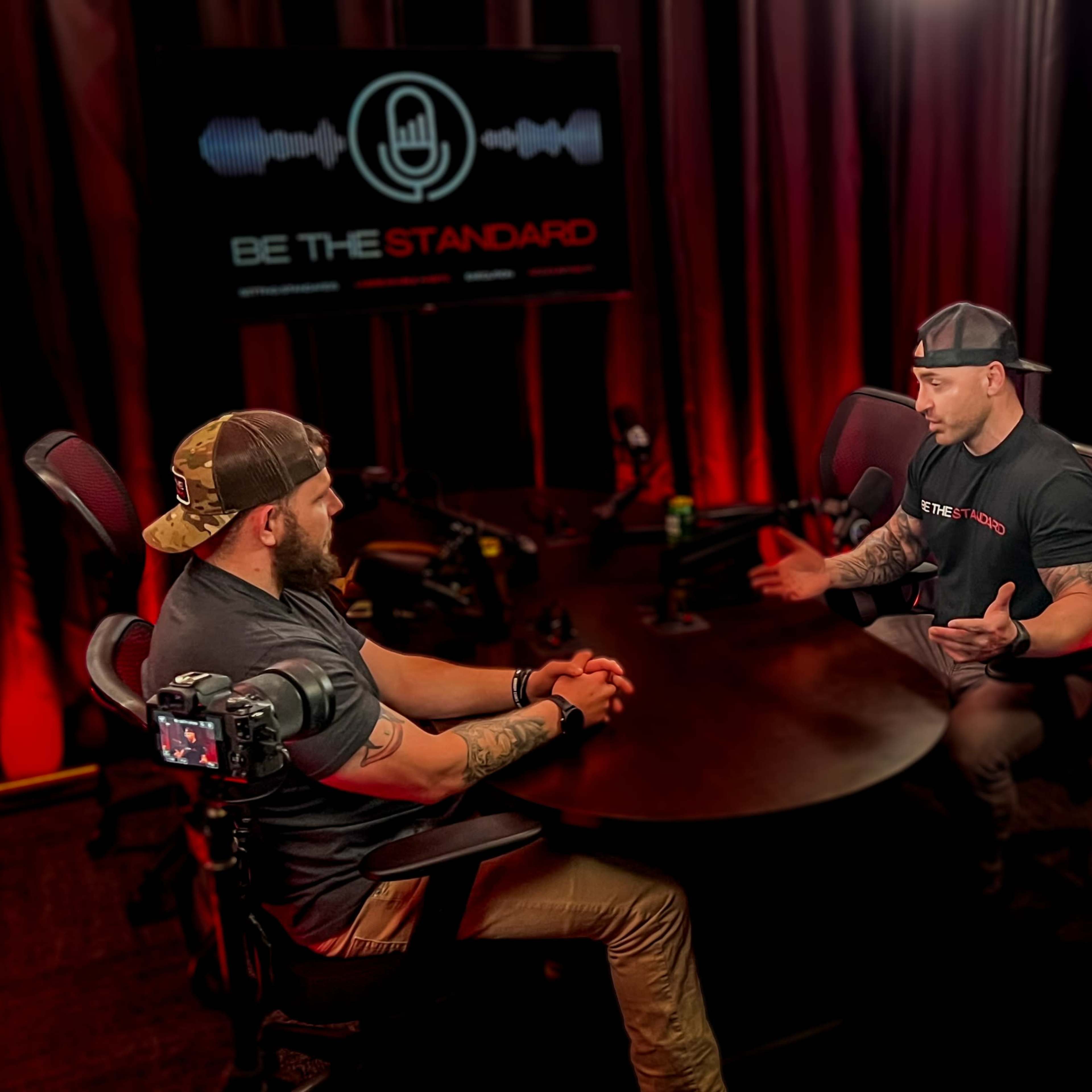 Two men are seated at a round table in a recording studio, discussing with microphones in front of them, while a screen behind them displays "BE THE STANDARD."