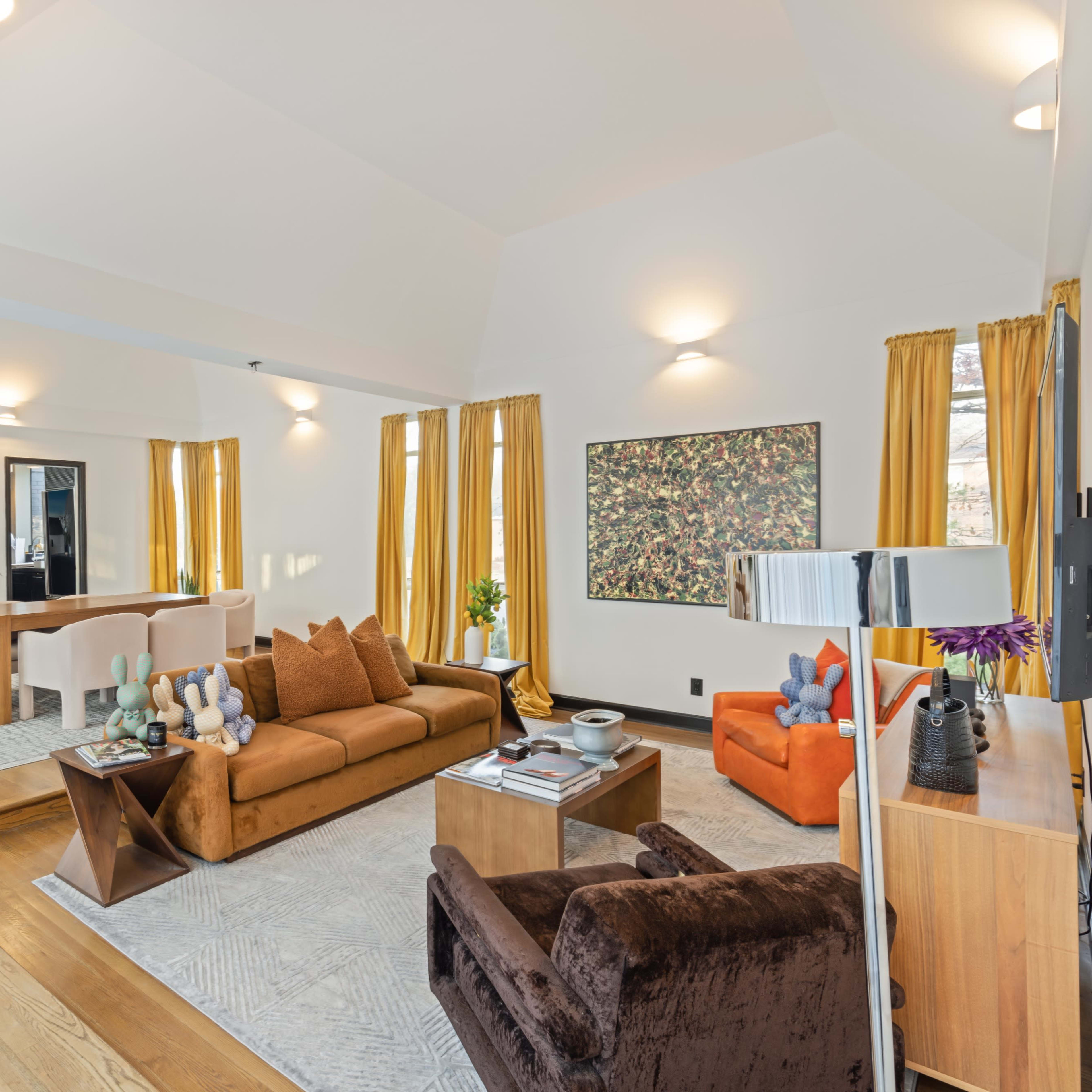 A modern open-concept living space featuring a brown sofa, an orange armchair, a dining area with a light table, and large windows with yellow curtains.