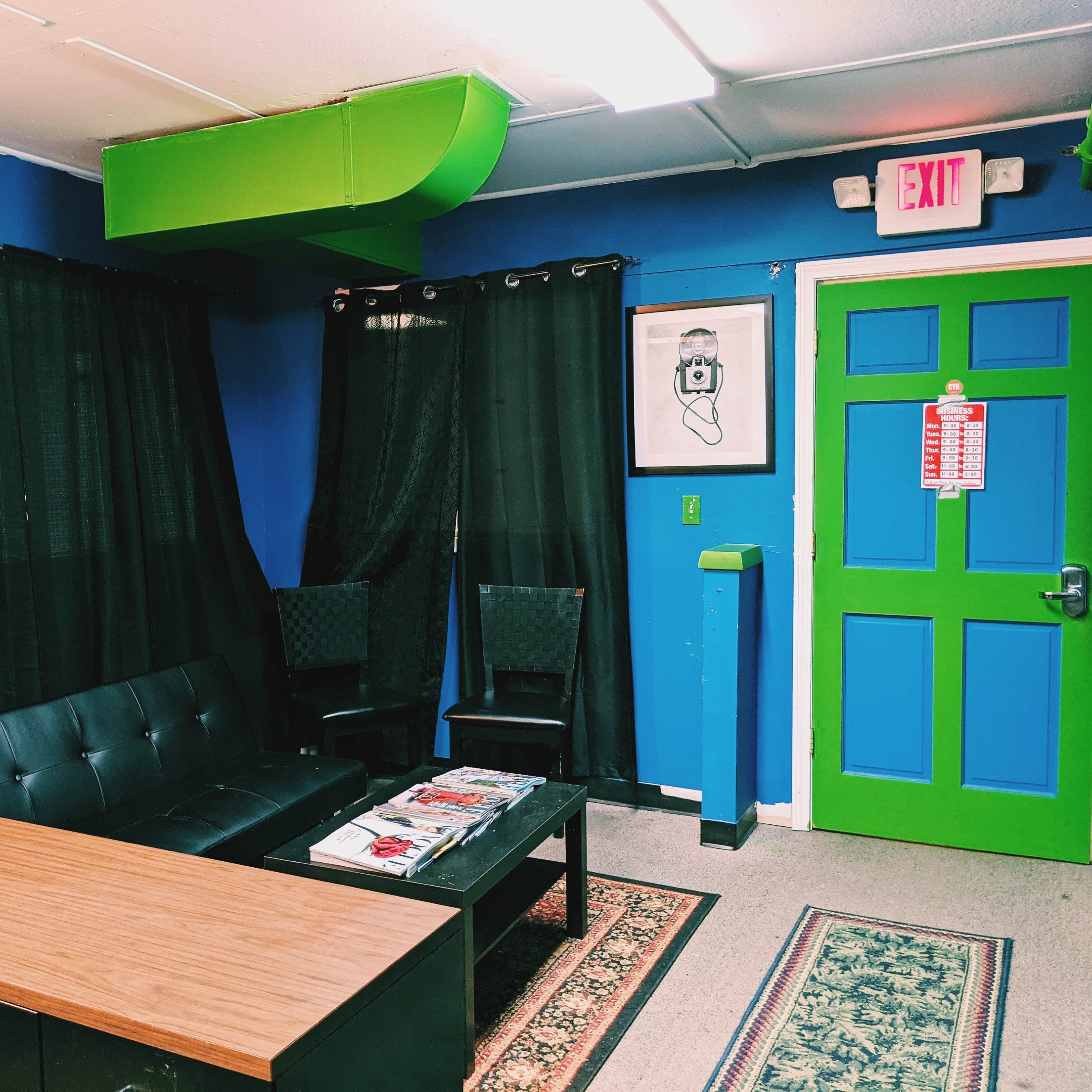 A small room with blue walls, a green ventilation duct, a dark sofa, two black chairs, a green door, and a shelf filled with various items.