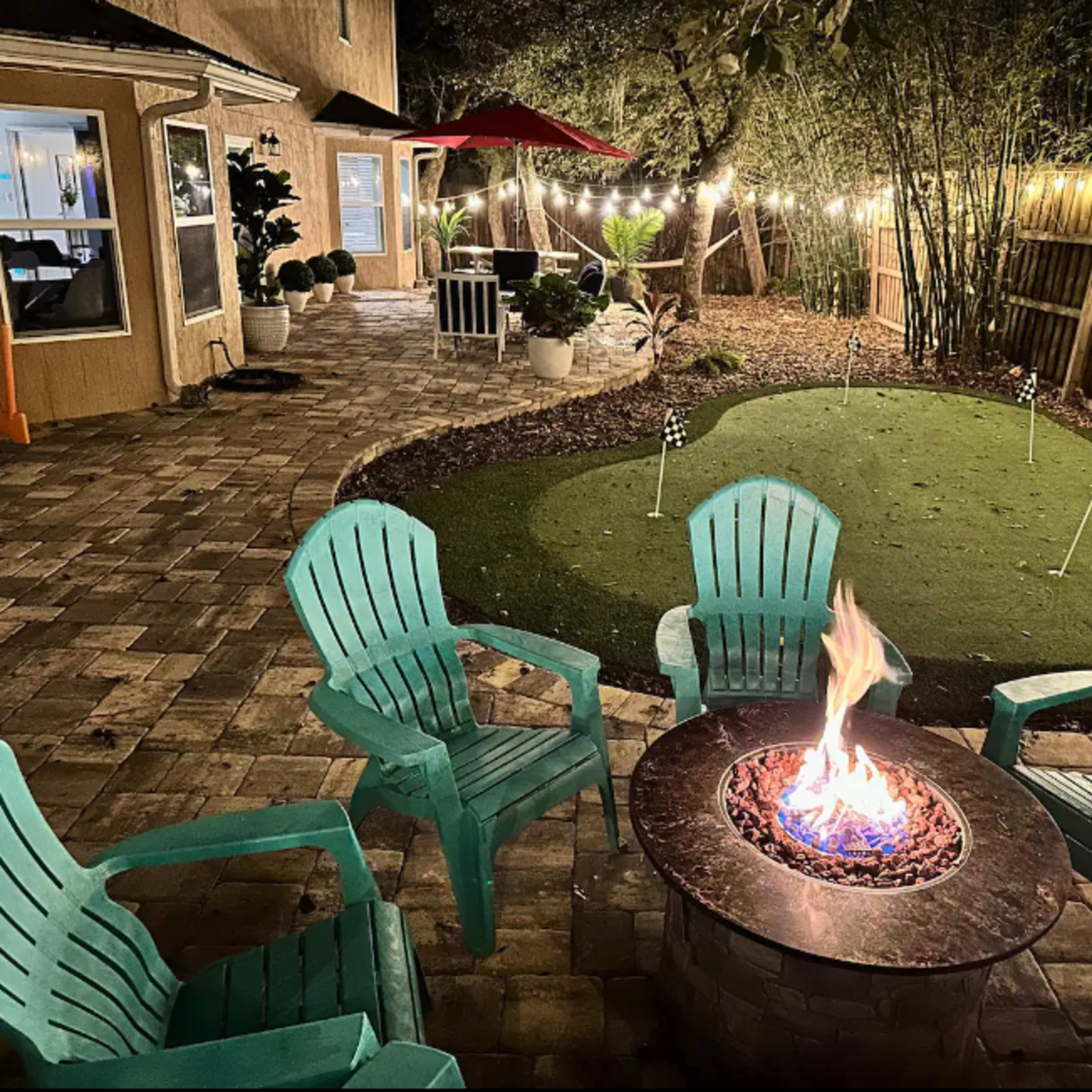 A stone fire pit surrounded by turquoise Adirondack chairs is located in a landscaped backyard with a putting green and string lights overhead.