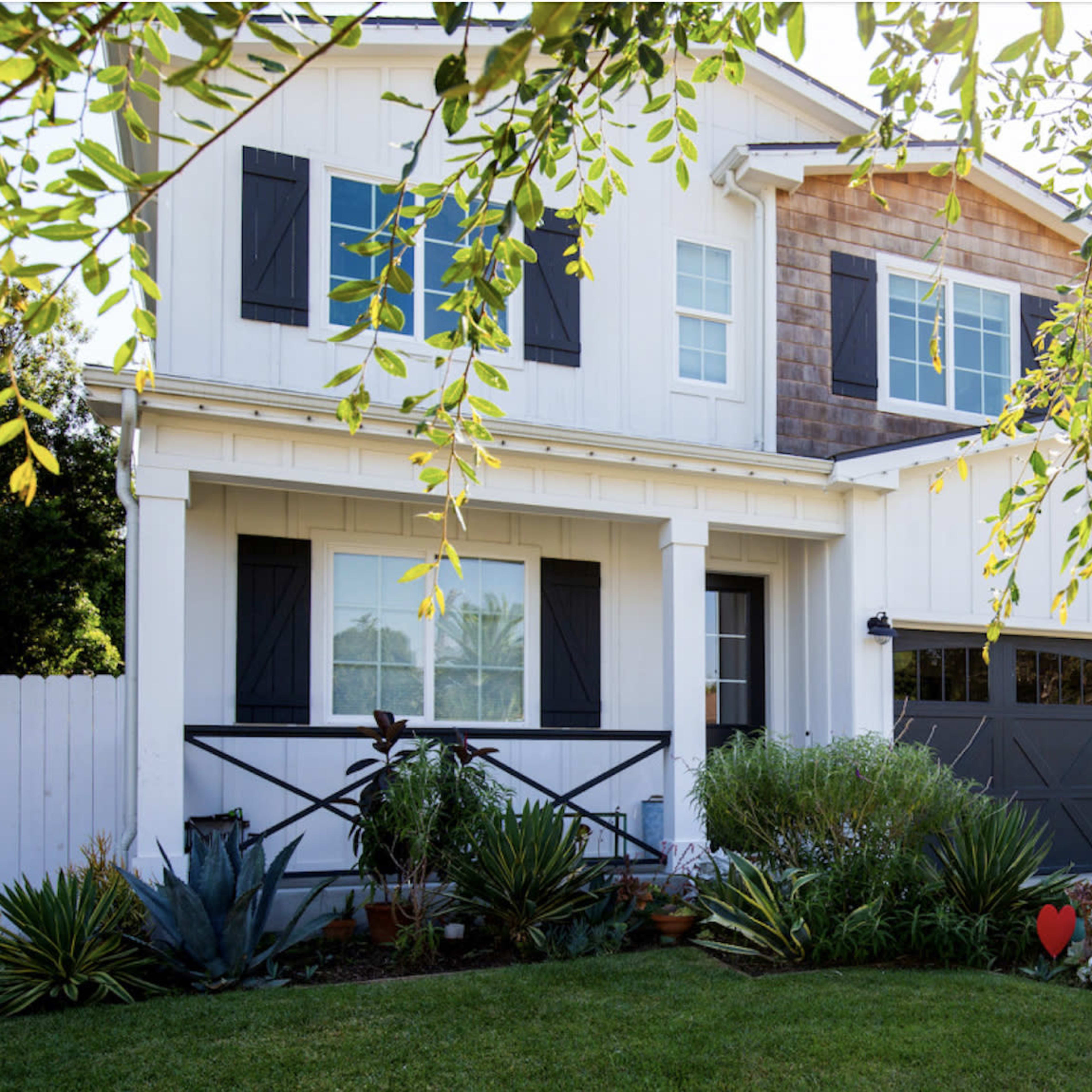 A two-story white house with black shutters and a brown shingle accent is surrounded by a well-maintained garden featuring various plants and a decorative heart.