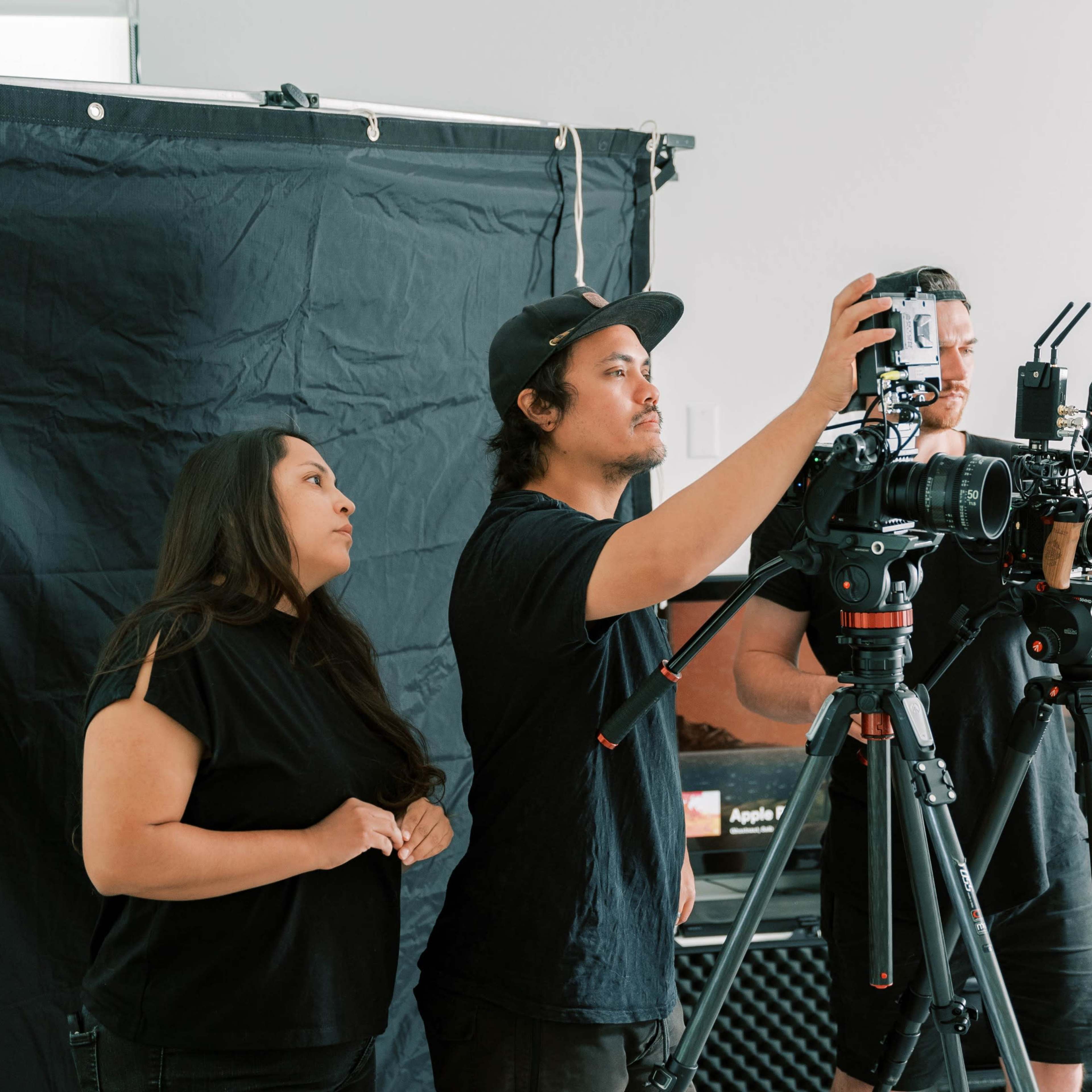 Three individuals operate cameras on tripods in a studio setting with a black backdrop.