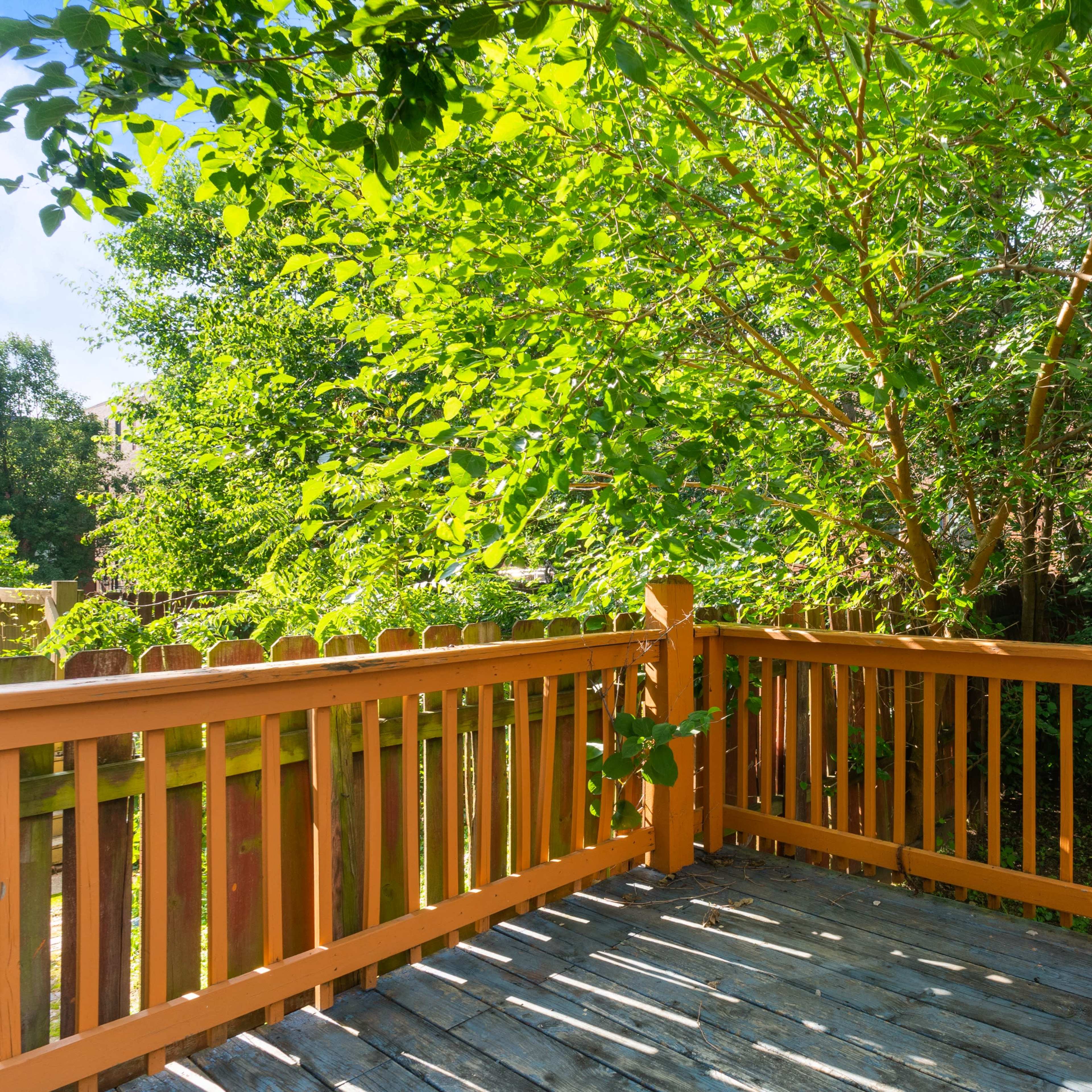 A wooden deck surrounded by lush green trees and a fenced yard is shown in the image.