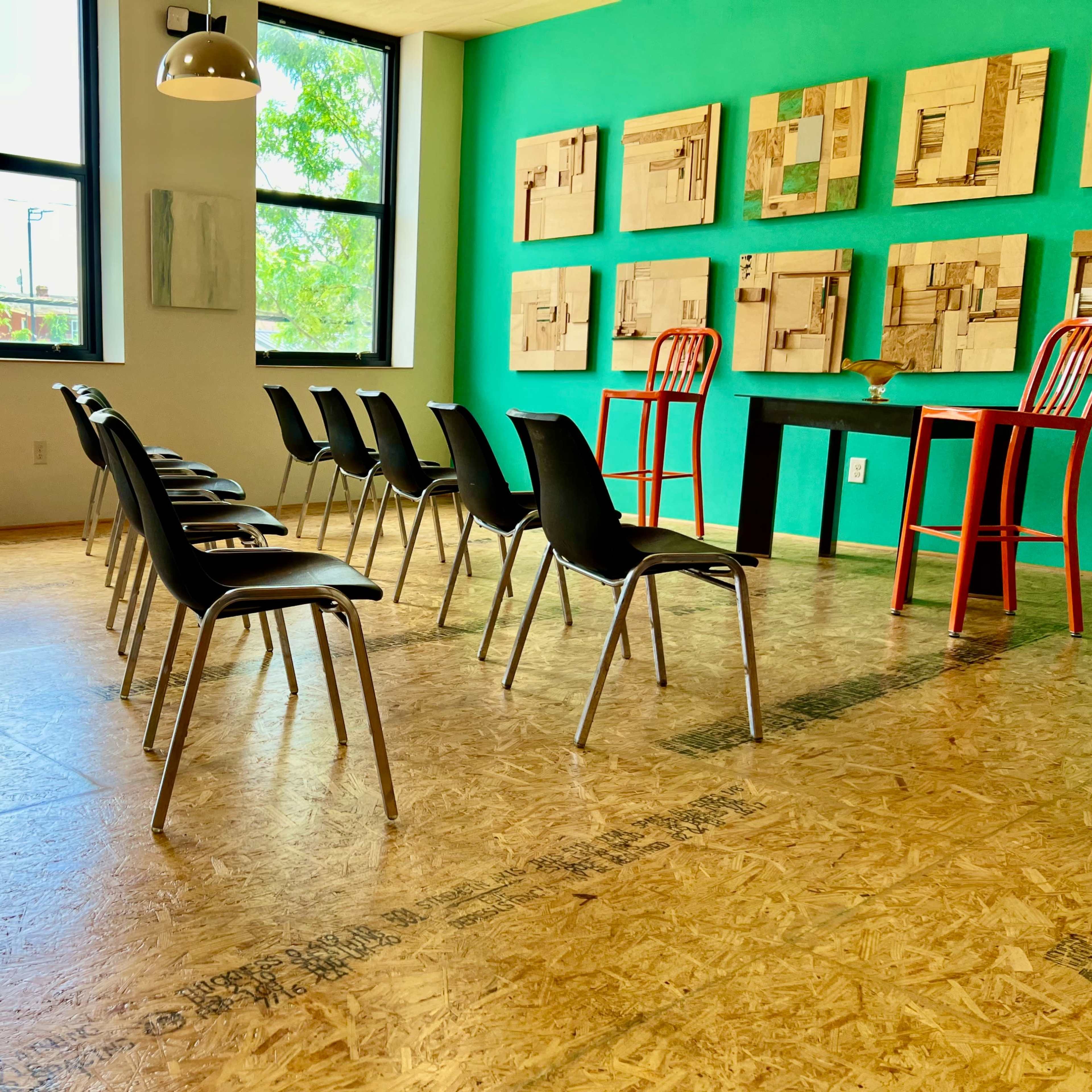 A room with several black chairs arranged in rows facing a green wall adorned with framed artwork, and two red chairs positioned at a small table.
