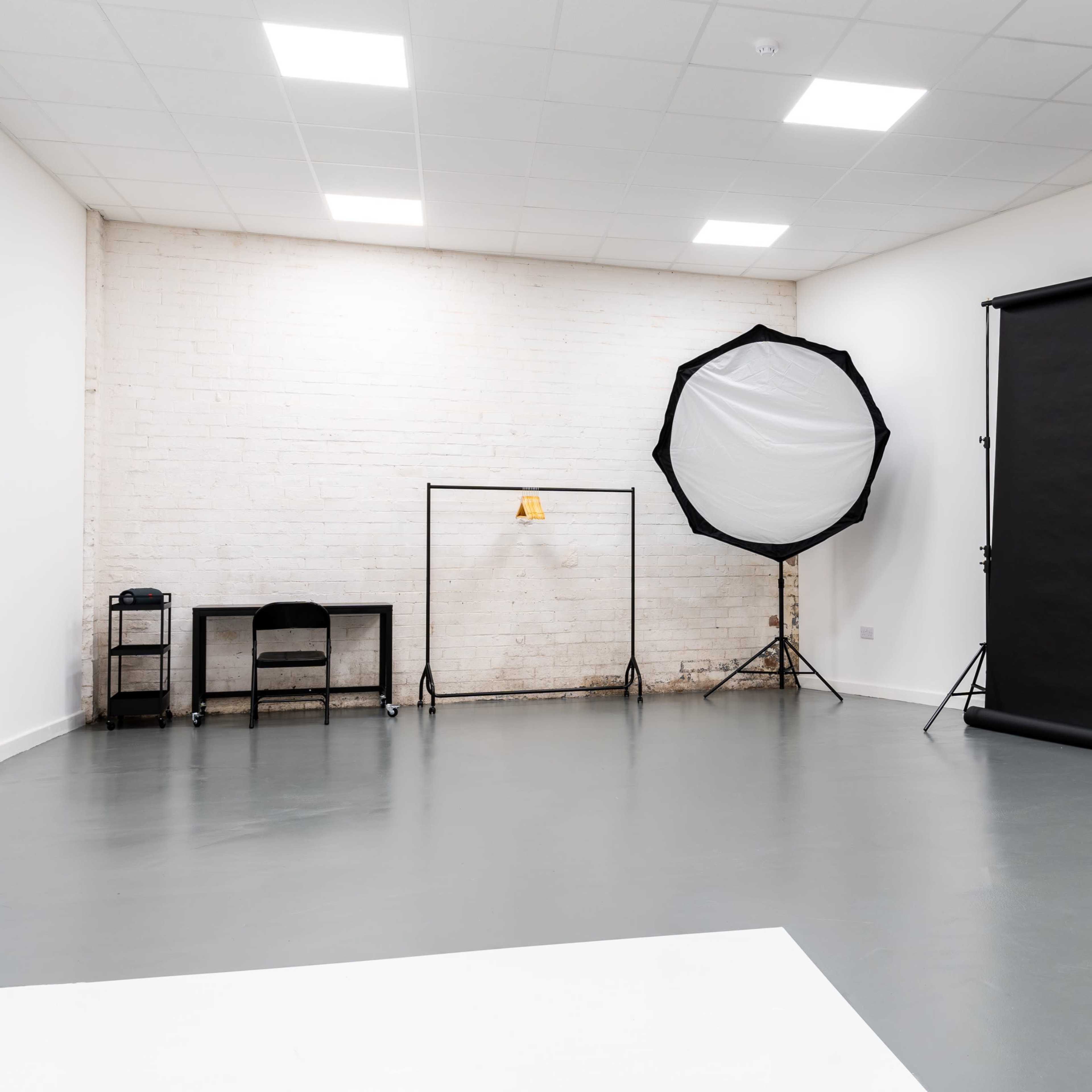A photography studio featuring a black backdrop, a large softbox light, and minimal furniture against a plain wall.