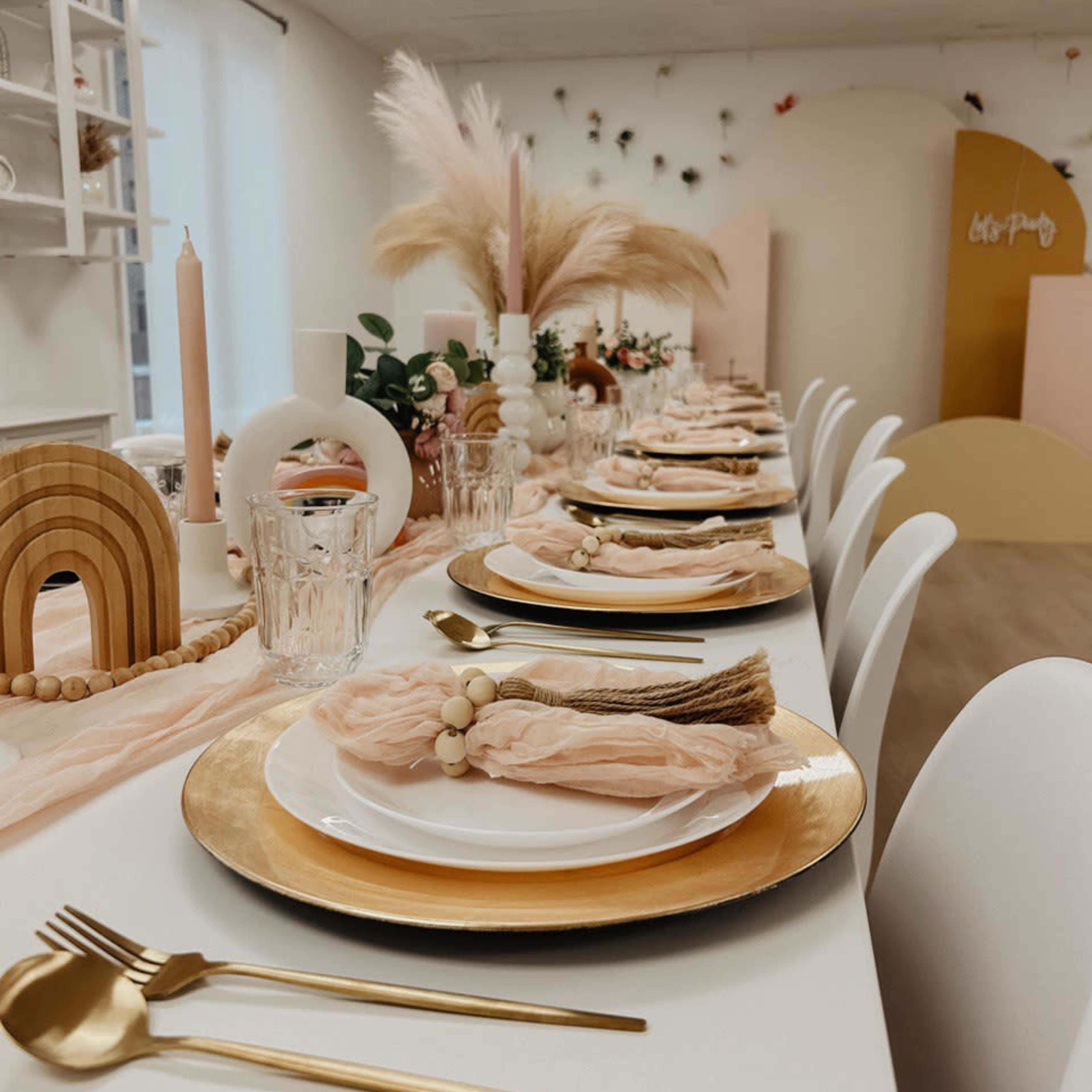 A long dining table is set with gold chargers, white plates, and blush napkins in an elegantly decorated room featuring pastel accents and decor.