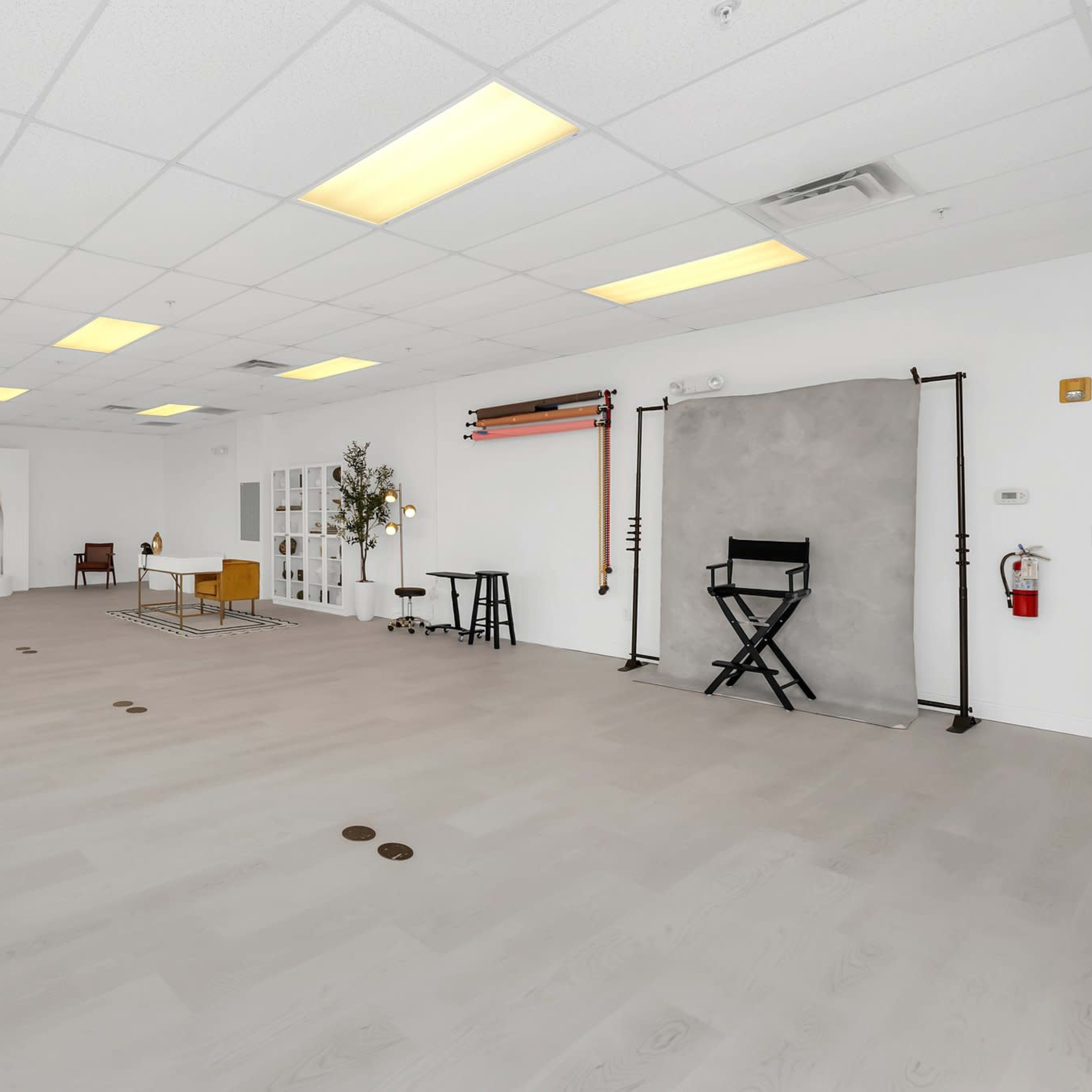 A spacious, minimalist photography studio featuring a light-colored wooden floor, various lighting equipment, and a neutral backdrop area.