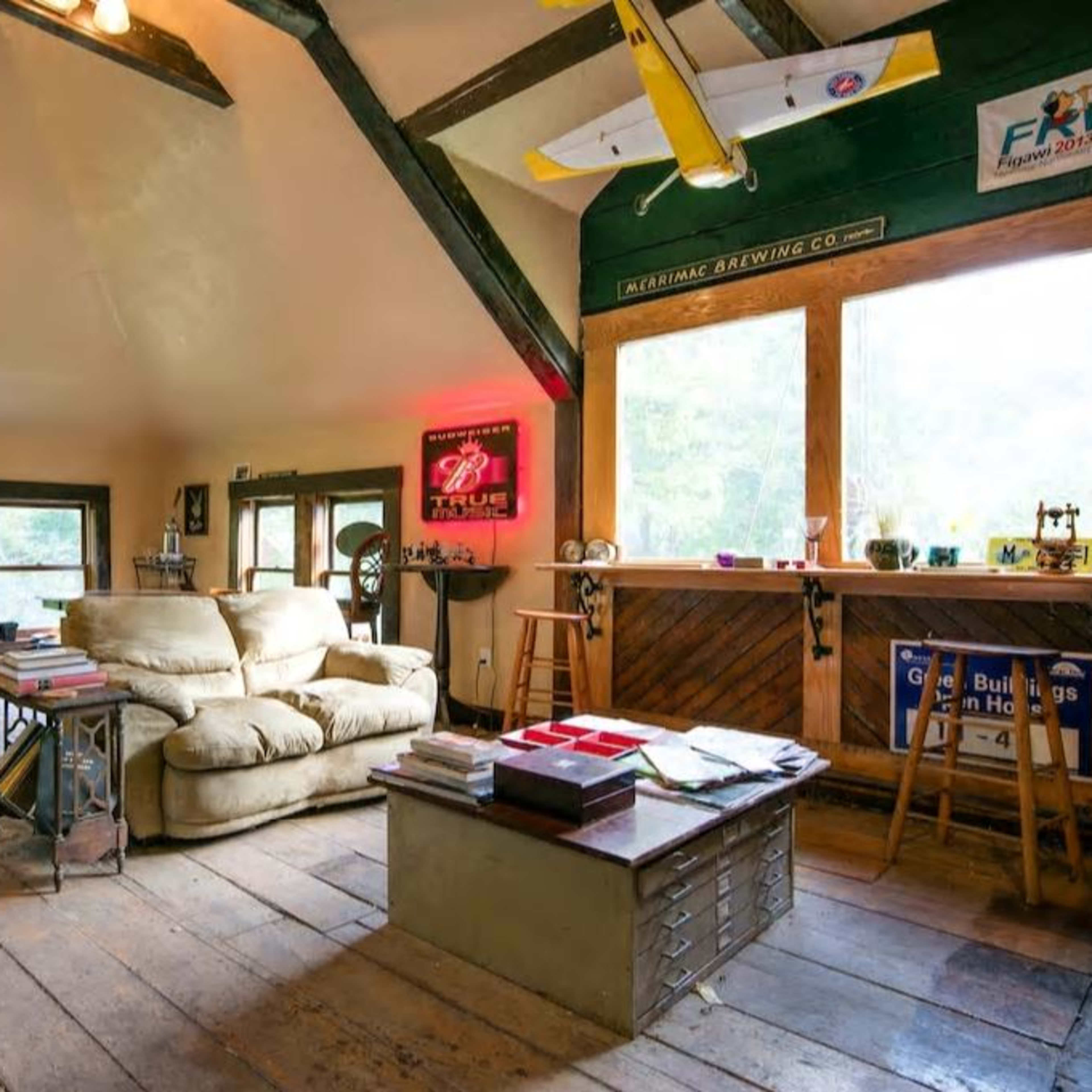 A cozy, rustic living space featuring a large couch, wooden dining table, and a bar area with windows overlooking a wooded view.