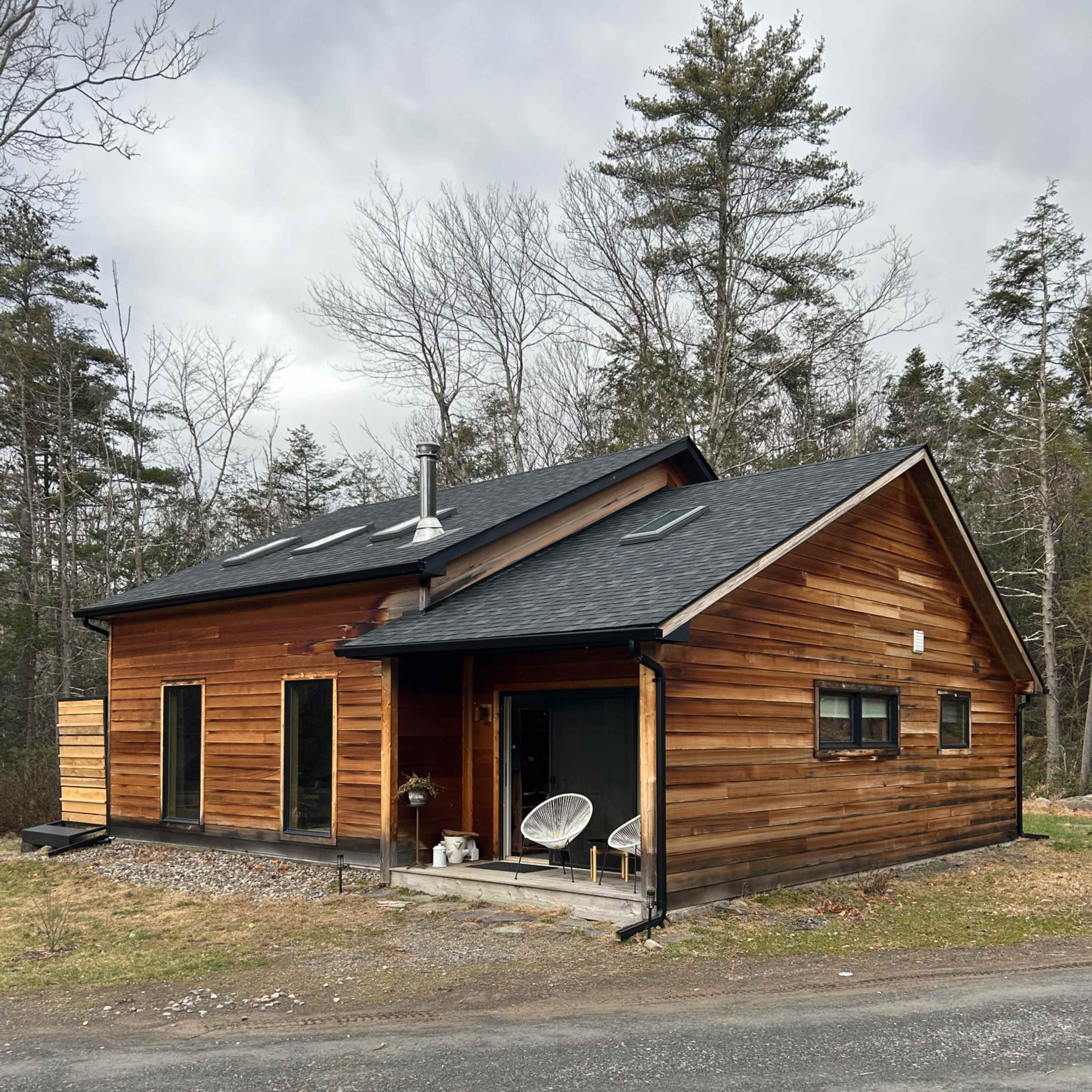 A modern wooden house with a sloped roof situated near a gravel road and surrounded by trees.